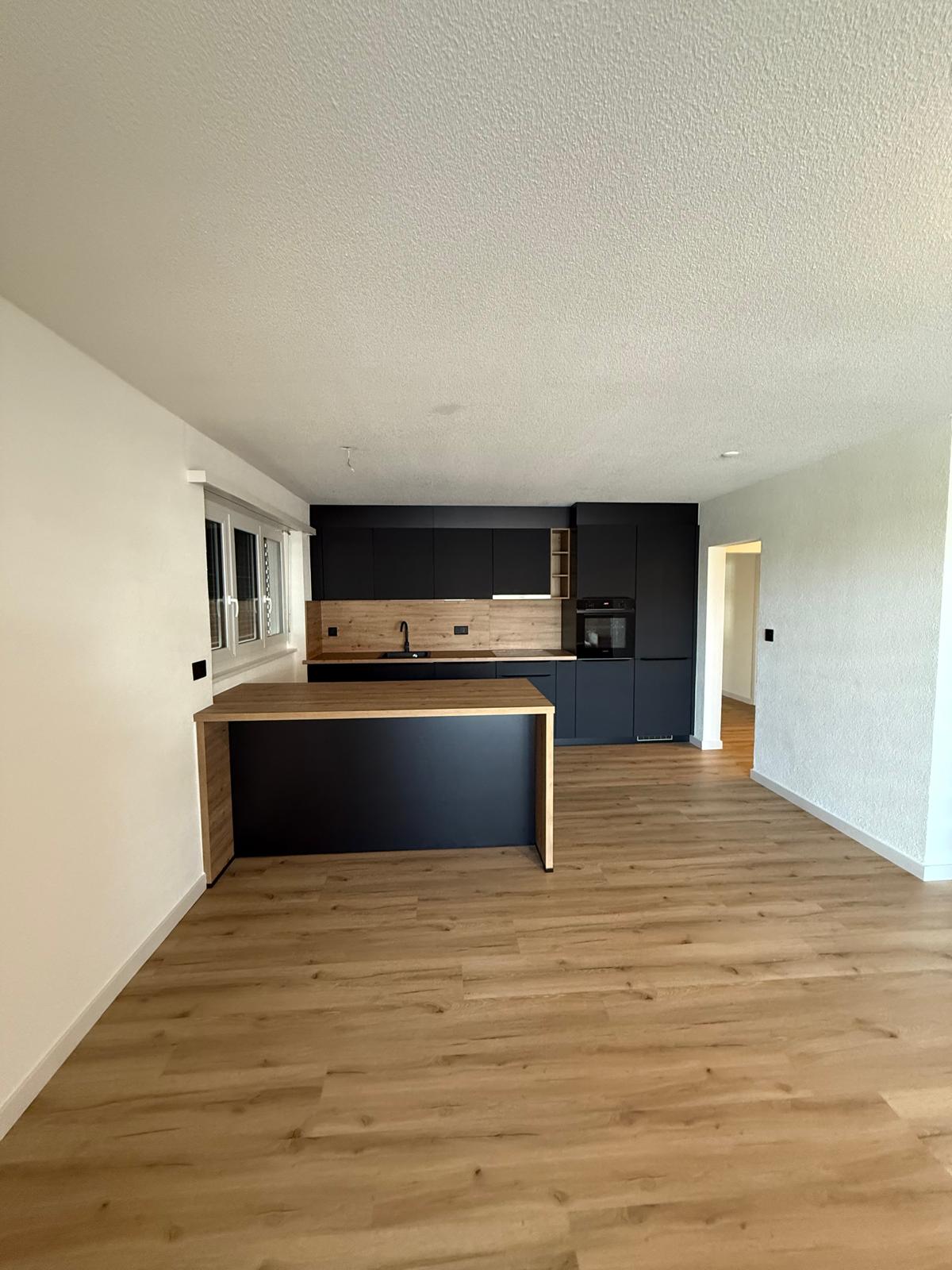 Empty modern kitchen with large island, black cabinets, white walls, and hardwood floors.