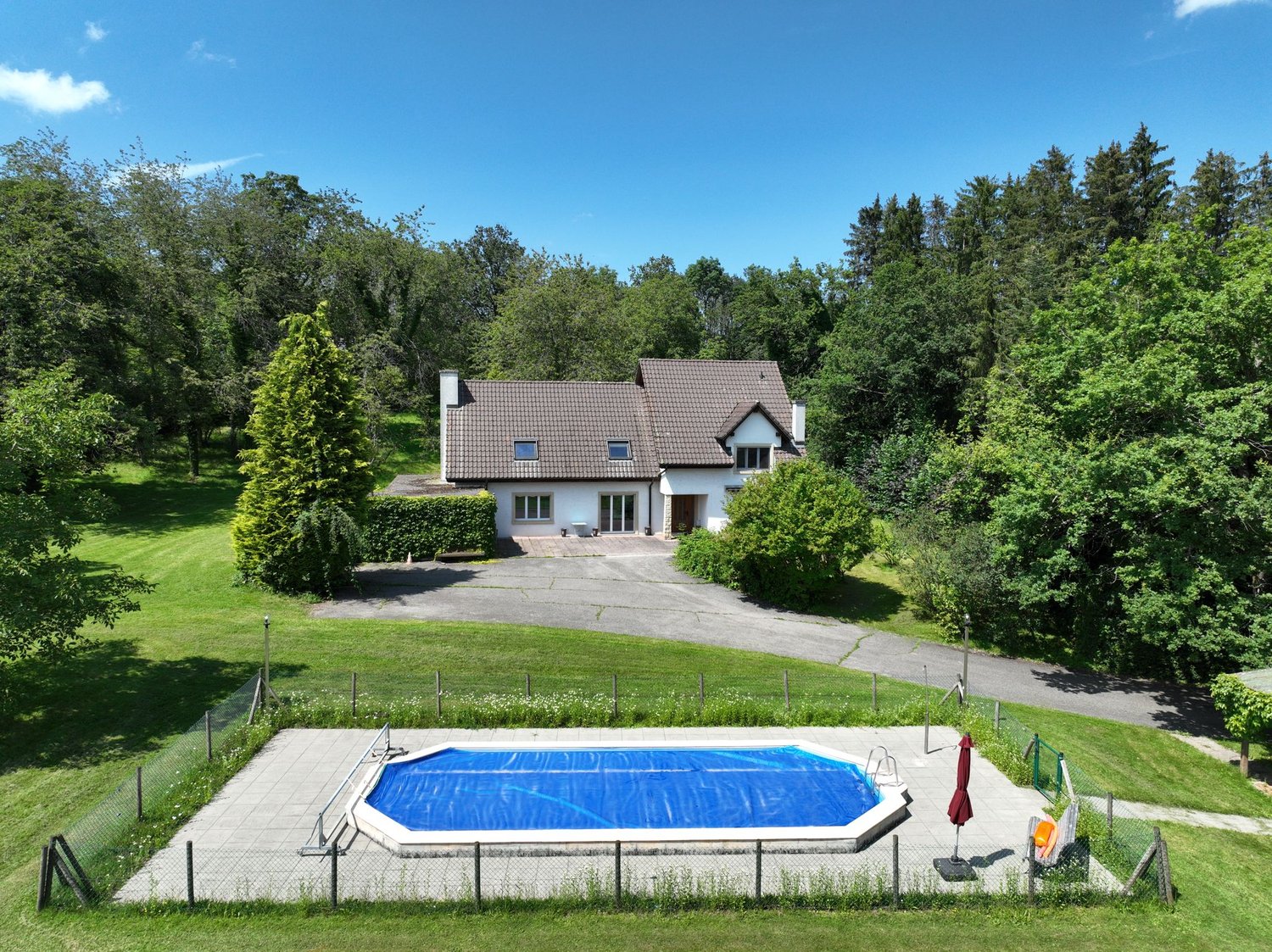 House with brown roof, white walls, 4 windows, driveway, swimming pool, green lawn, hedges, trees
