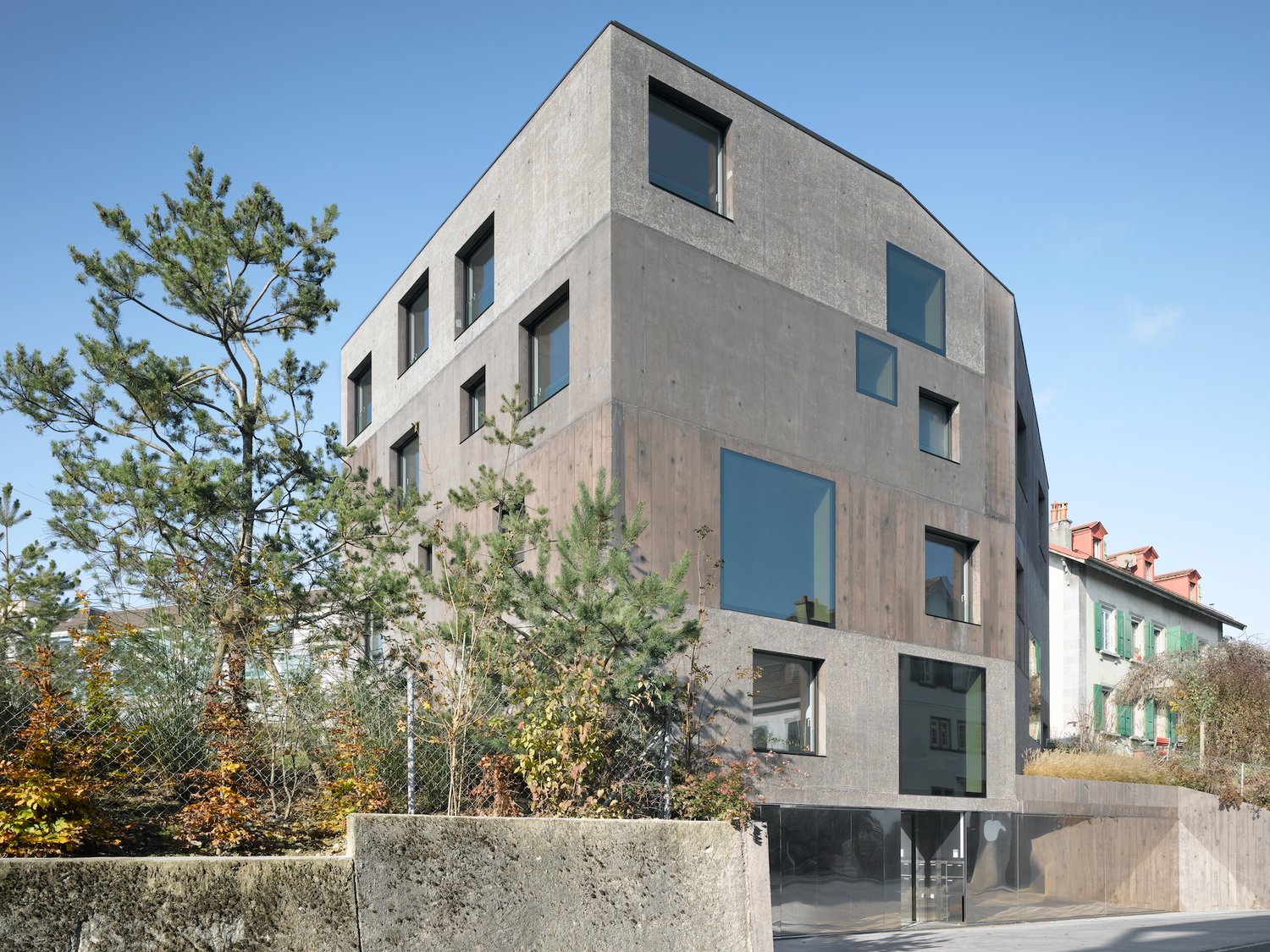 A modern, multi-story concrete building with large windows and a minimalist design. The building is surrounded by trees and other residential structures.