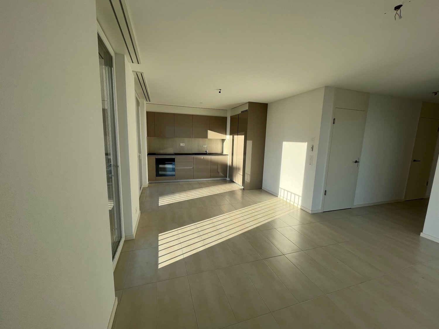 empty living room with kitchen visible, tiled floor, sliding glass door leading to a balcony, wooden cabinet doors
