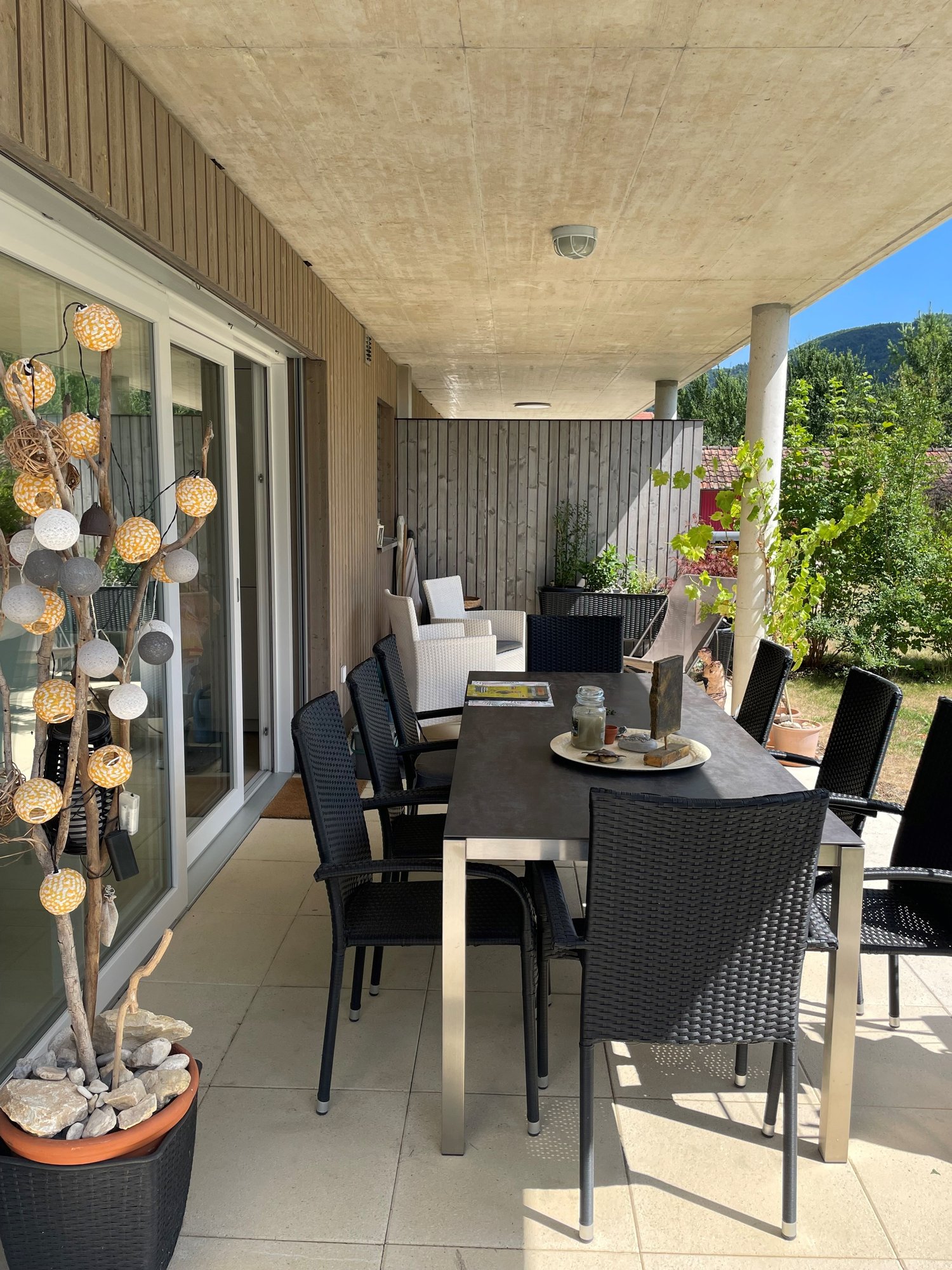 Covered outdoor patio with a dining table and chairs, wicker furniture, decorative lighting, and potted plants. The patio has a concrete floor and is surrounded by a wooden fence with a view of the surrounding greenery.