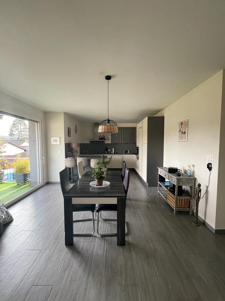 The image shows an open-plan living room and kitchen area. The room has a large dining table with chairs, a kitchen with dark cabinets and appliances, and a sliding glass door leading to a balcony or terrace. The floor is covered in a dark wood-like tile,