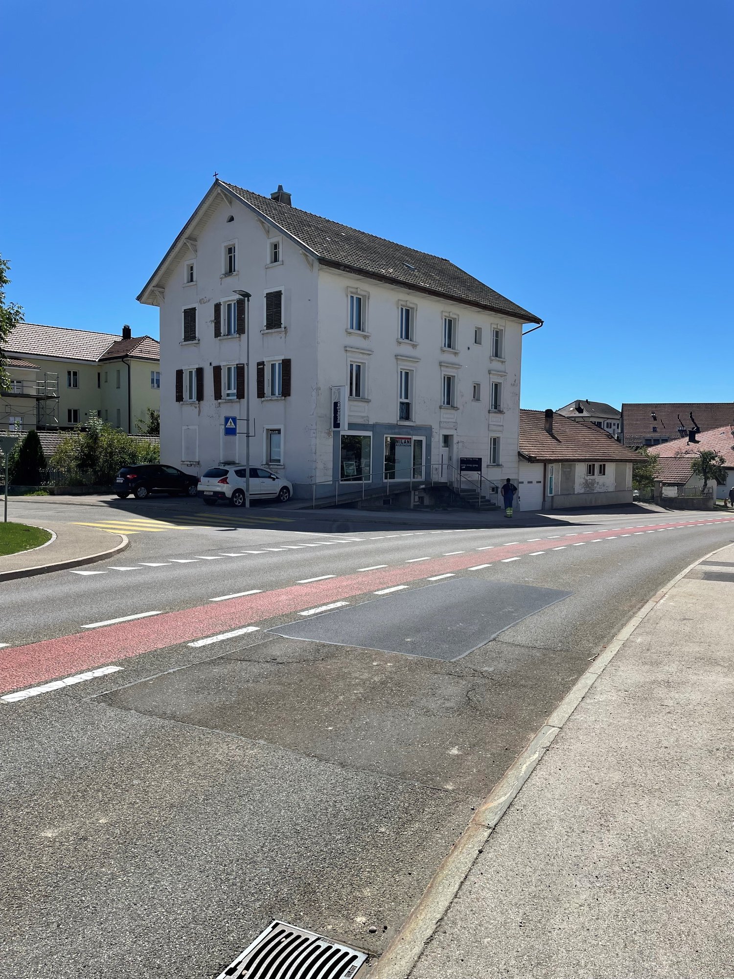 Large white house, multiple windows, red roofs, parked cars in front, two other houses behind the road
