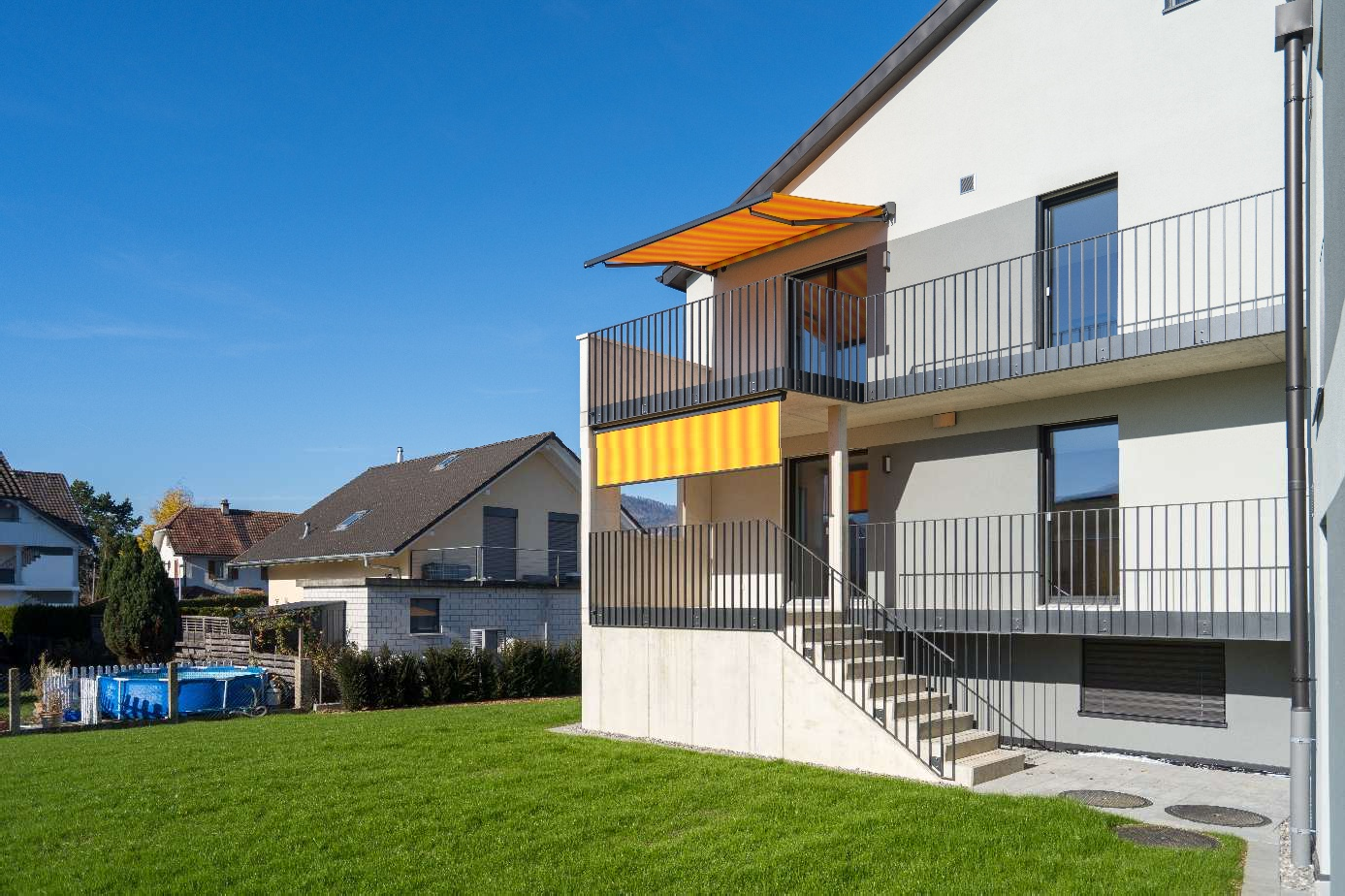 2 story house, modern architecture, white exterior, orange and white awning, black railings, two balconies, concrete ground floor, green lawn