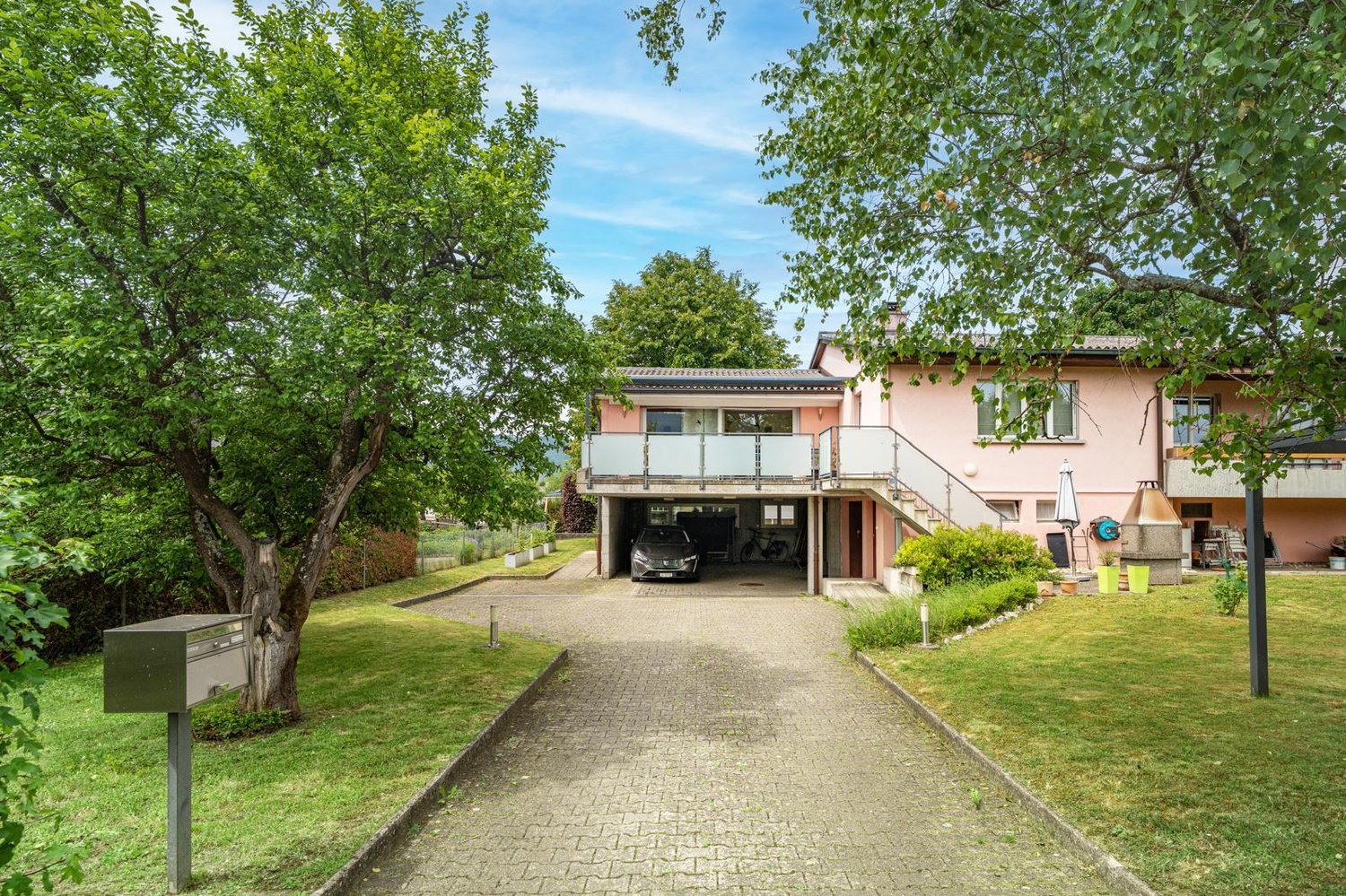 Two-story house with a pink exterior, surrounded by lush greenery and trees. The house has a balcony on the upper level and a garage with a parking space underneath. The property has a paved walkway leading to the entrance and a well-maintained lawn.