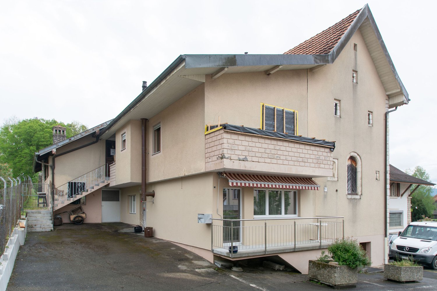 2-story house, beige exterior, brick base, staircase, balcony, awning, large driveway, plants, fence, parked car