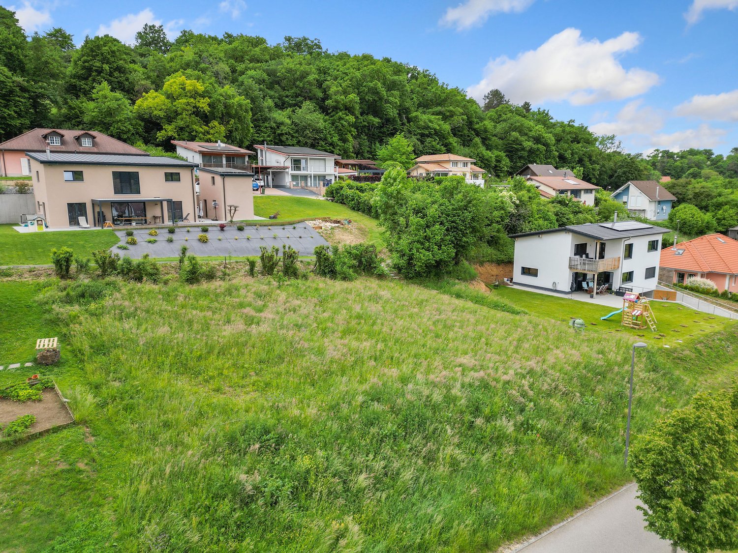 The image shows a residential area with several multi-story houses and apartments nestled in a lush, green landscape. The buildings have balconies and terraces, and the surrounding area is covered in grass and trees. The scene appears to be in a suburban