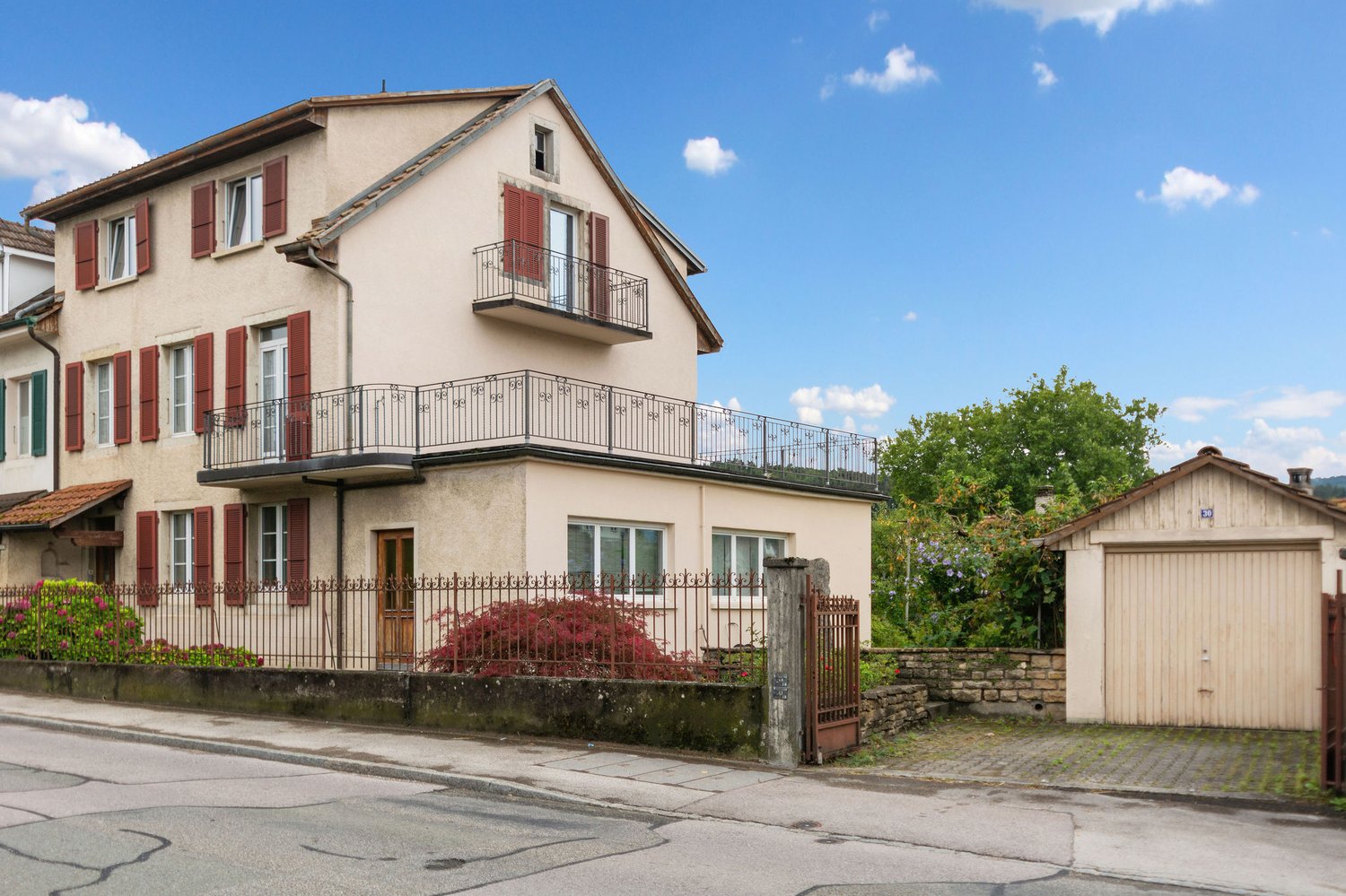 2 story house, beige exterior, red shutters, iron railing on balcony, wooden front door, garage at side, black gate