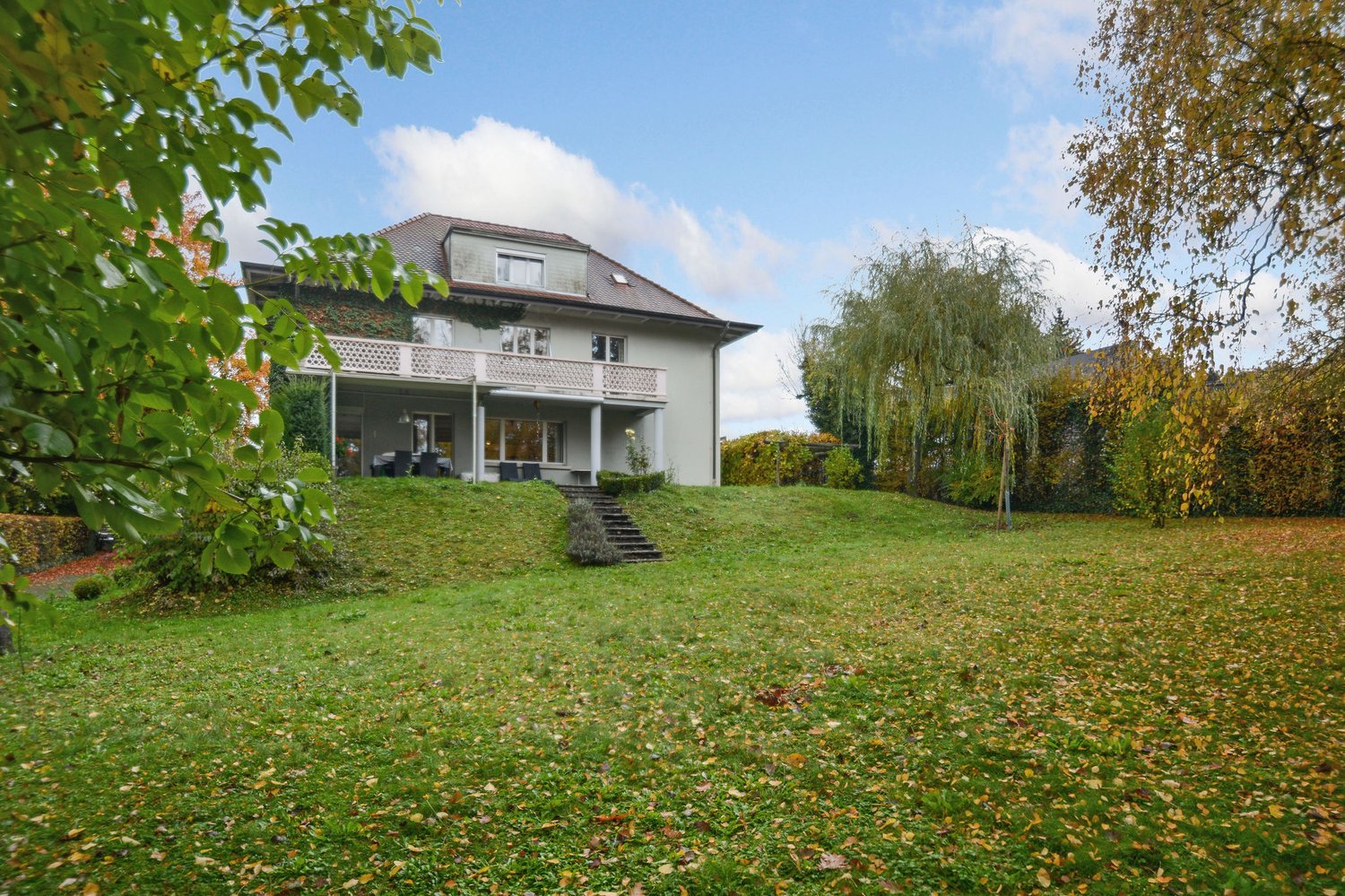 A two-story house with a large lawn in front. The house has a balcony with white railings on the second floor. The house is on an elevated land.