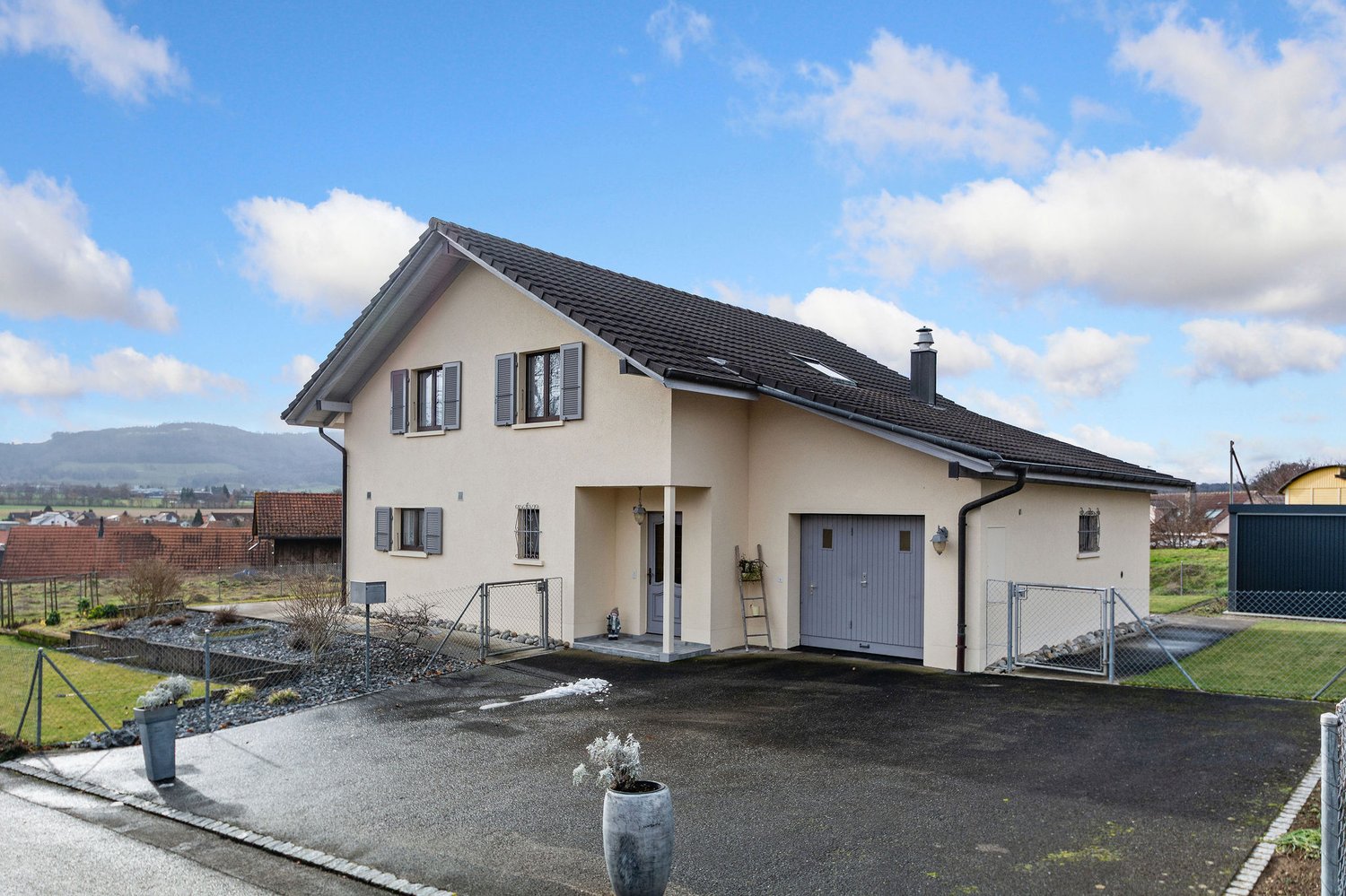 Modern house, beige exterior, black roof, large windows, two garages, driveway, fenced garden, mountains in the distance