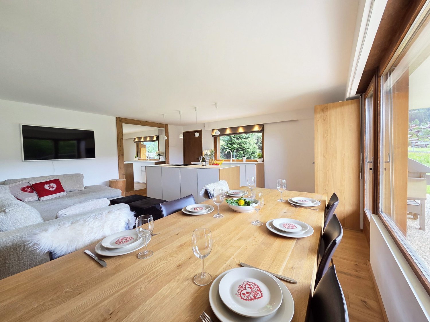 Spacious open-plan living area with a large wooden dining table, modern kitchen with white cabinets, and a cozy seating area with a gray sofa. The room has large windows providing a view of the outdoor scenery.