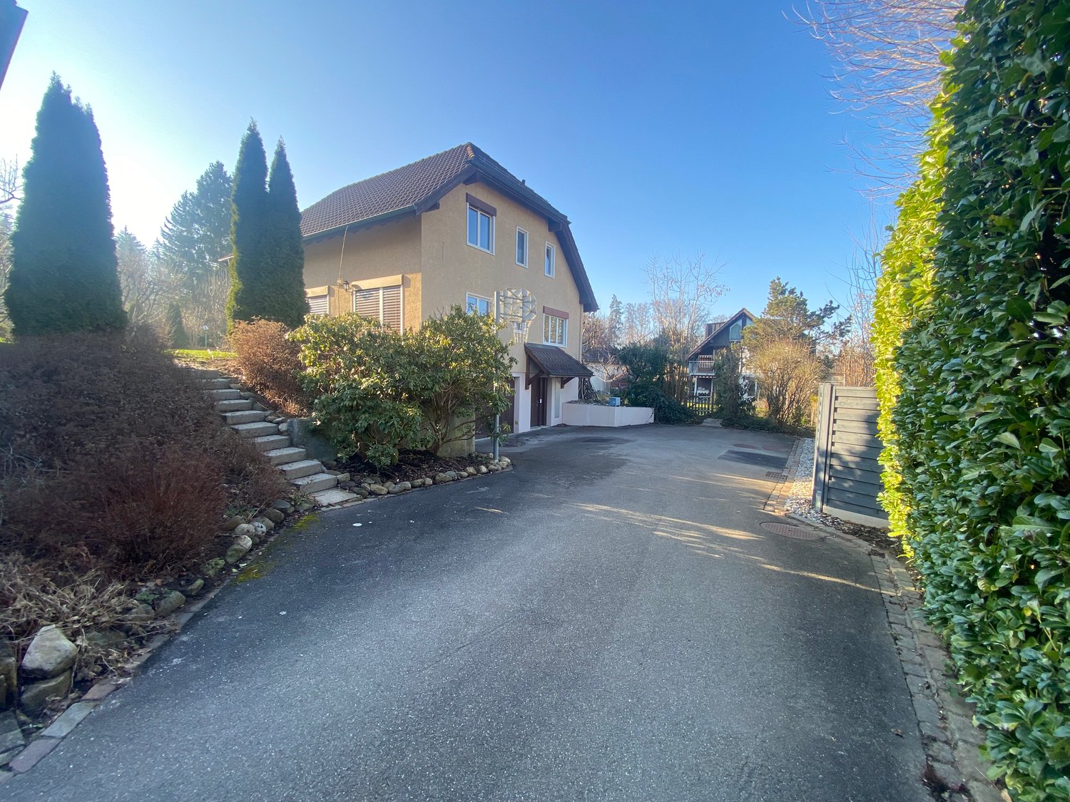 2 story house, beige walls, brown roof, paved driveway, front stairs, hedges, side gate