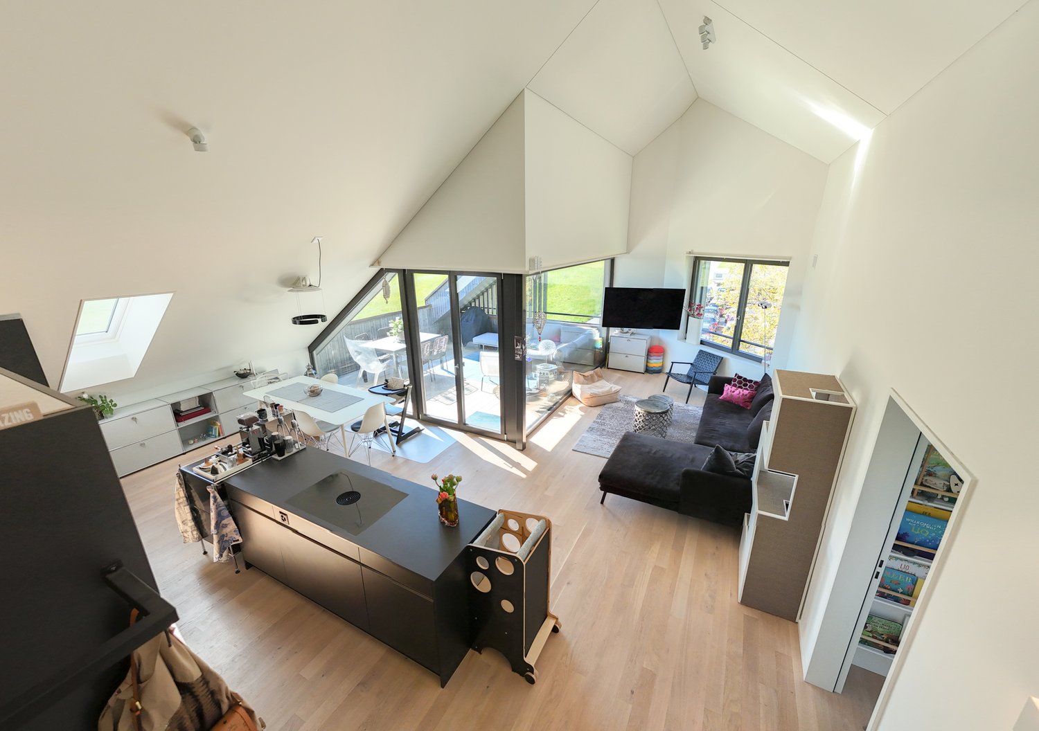 Modern open plan living, dining and kitchen area with large windows, central island, black countertops, white cabinets, kitchen island with modern appliances, black couch, coffee table, armchair, fireplace, shelves, television