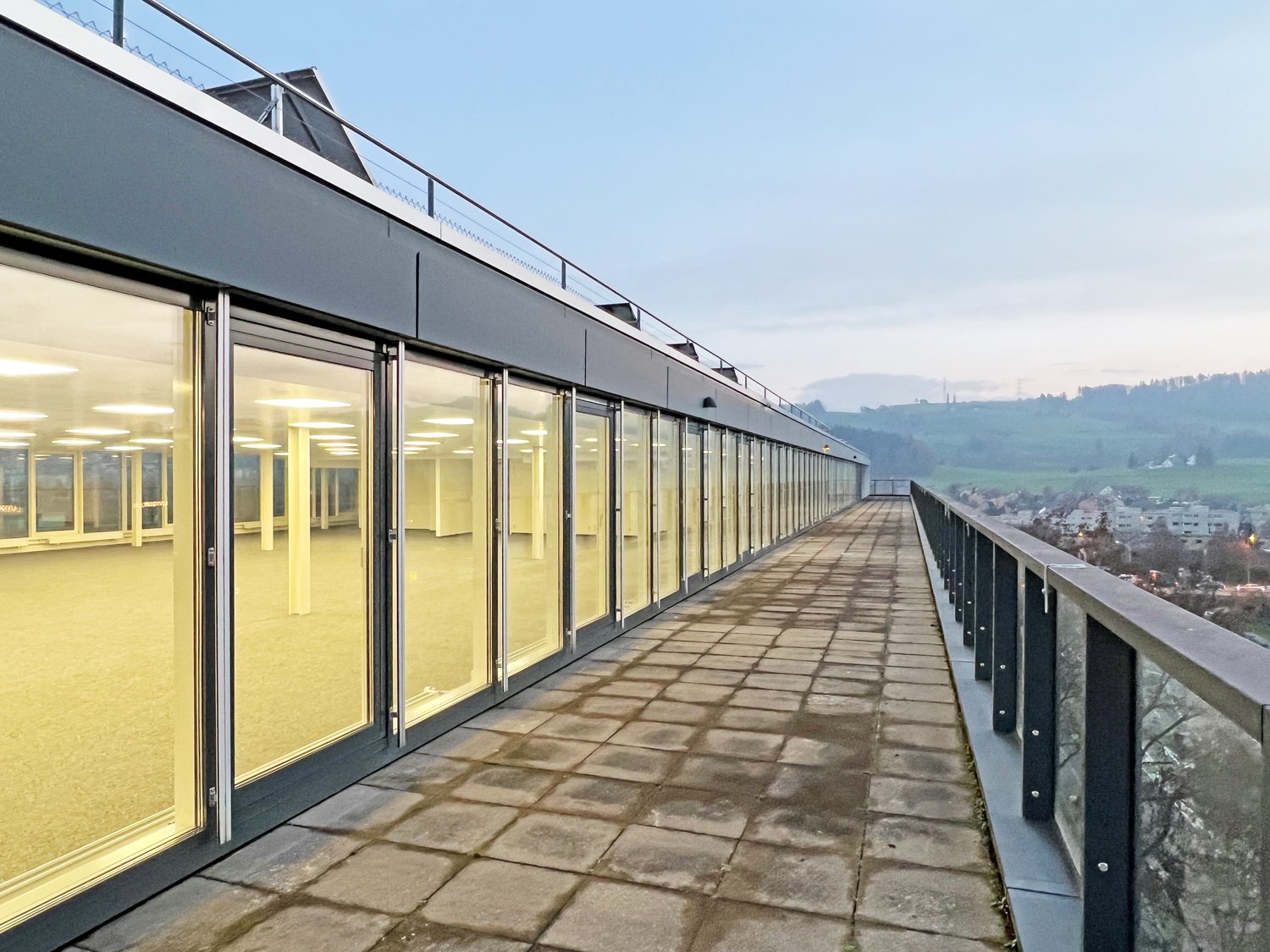A multi-story commercial building with a long balcony or terrace running along the exterior. The building has large windows and a modern, minimalist design. The balcony offers a scenic view of the surrounding landscape and cityscape.