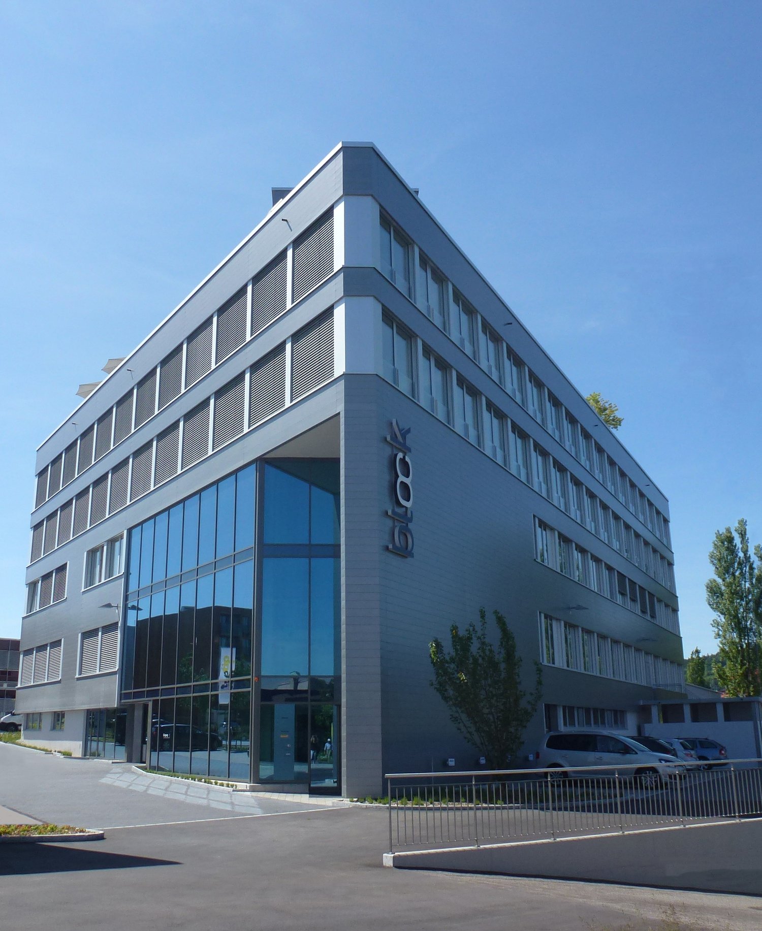 A modern, multi-story office building with a sleek, angular design. The building has large windows and a prominent logo on the facade. There is a parking area in front of the building.