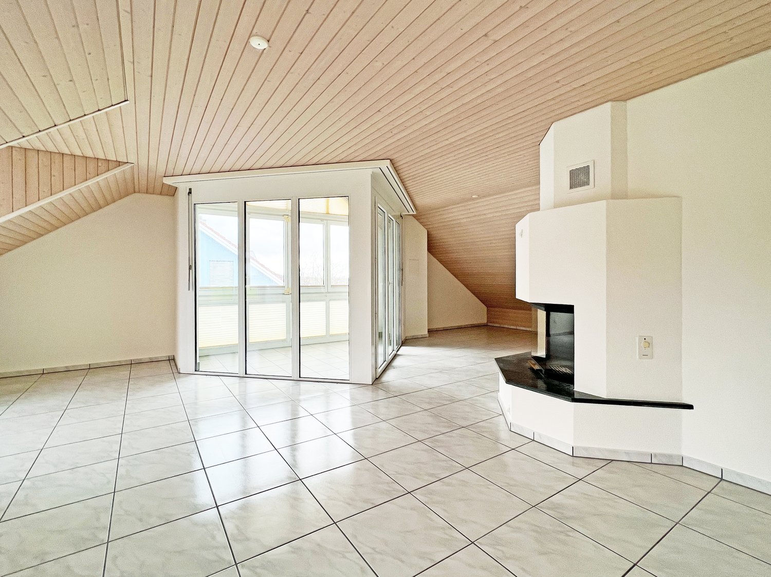 Spacious attic living room with wood-paneled ceiling, large windows, tiled flooring, and a fireplace