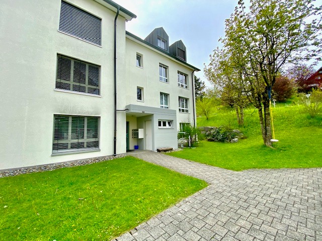 Two story apartment building with exterior brick wall, large windows, pathway leading to entrance, green lawn, small trees, and shrubs