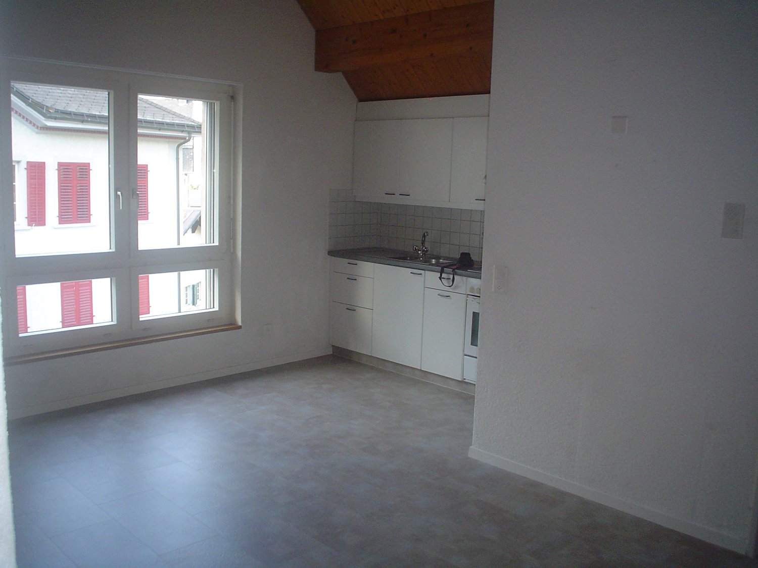 Empty kitchen, tiled floor, wooden ceiling, double glazed window, white cabinets, stainless steel sink, tile backsplash, black cord on counter