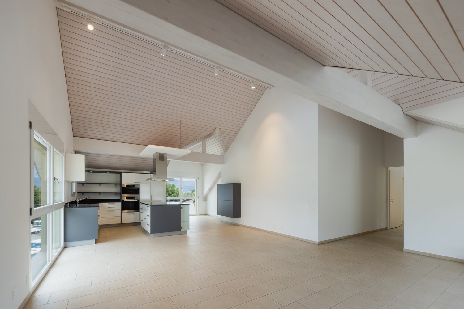 Spacious open-plan living area with vaulted ceiling, wood paneled ceiling, modern kitchen with built-in appliances including a dishwasher, and large windows providing natural light