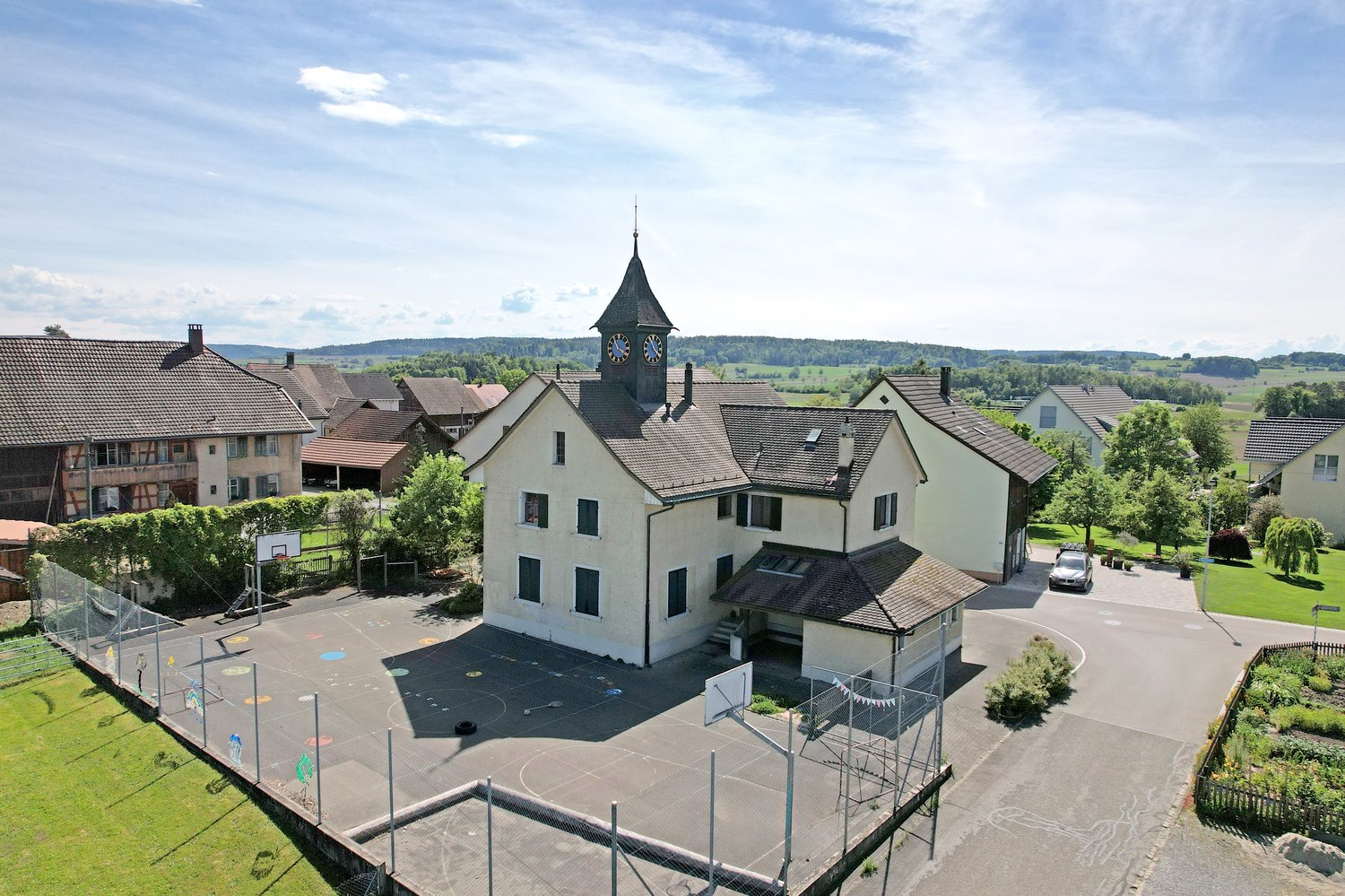 Aerial view of a large white house with a tower and a clock, surrounded by trees and a basketball court.
