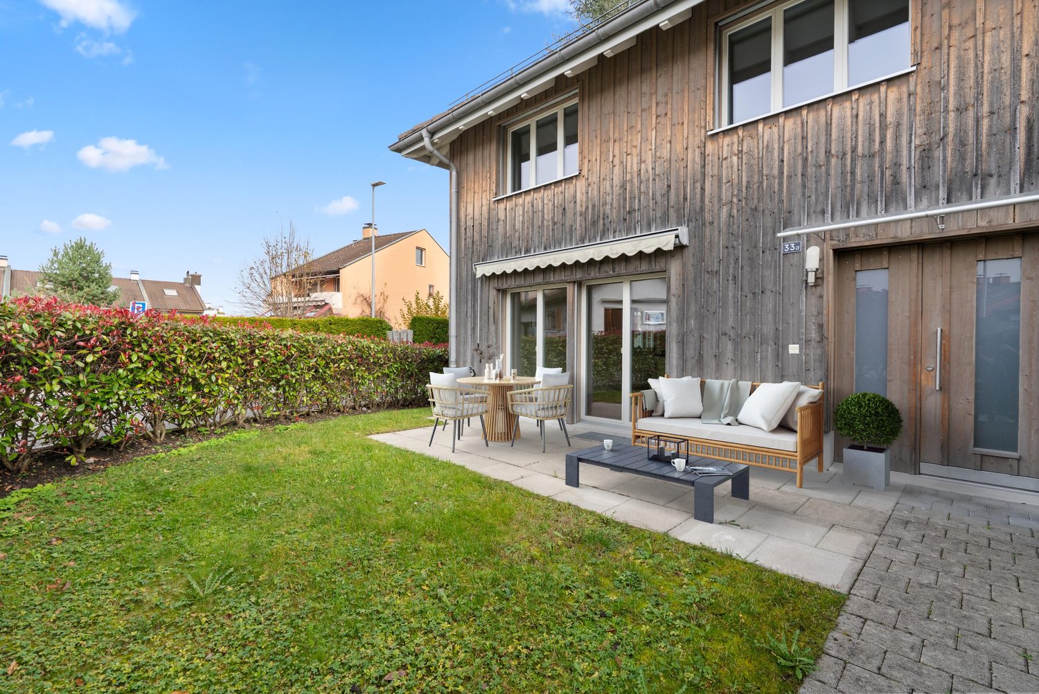 a brown wooden house with a patio, table, chairs, sofa, outdoor table, and a well-kept garden with a brick pathway