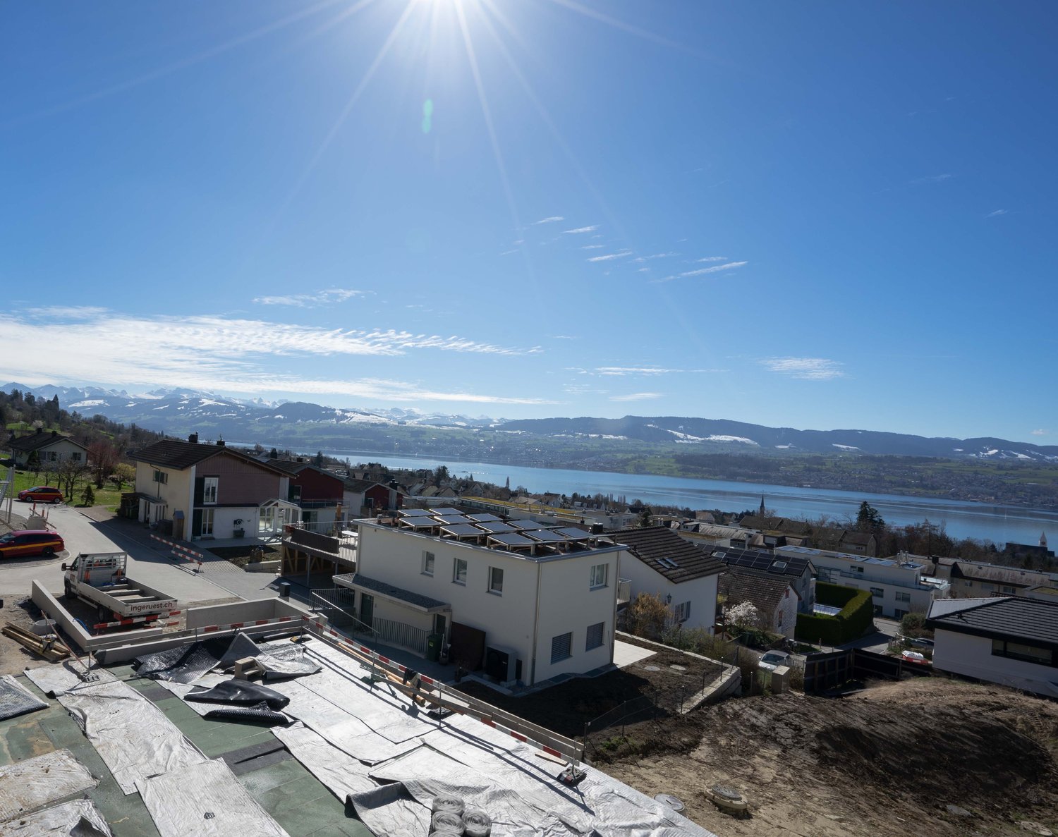 House under construction, solar panels on the roof, surrounded by houses, mountains, and a lake in the background