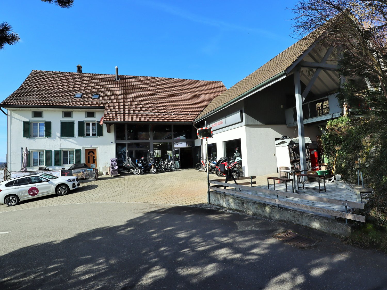 Two buildings, one with brown roof, white walls, several windows with green shutters, garage with parked motorcycles, and another building with an awning and a table outside. A car is parked in front of the first building.