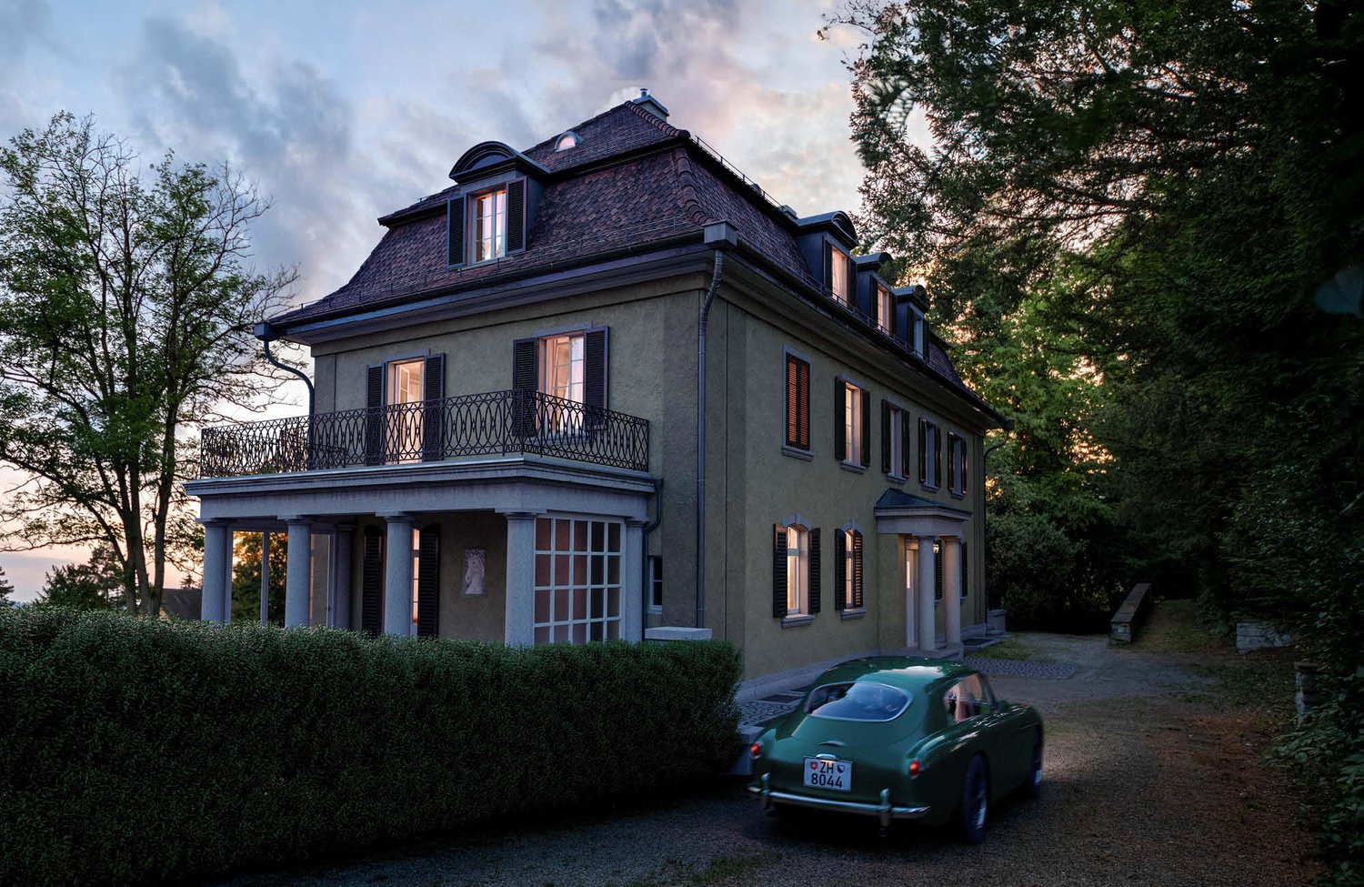 Two-story mansion with a slate roof, dormer windows, and a balcony. The exterior is painted in a dark gray color. There is a vintage car parked in the driveway in front of the house. The house is surrounded by trees and greenery, creating a picturesque se