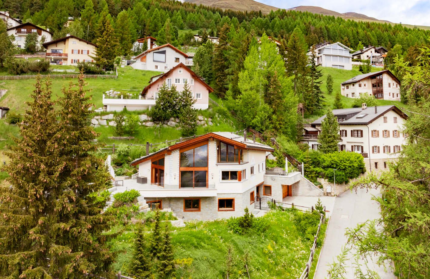 Scenic mountain village with traditional Swiss-style houses, surrounded by lush green forests and hills. The image shows a residential area with several multi-story houses, some with balconies or terraces, nestled in the picturesque alpine landscape.