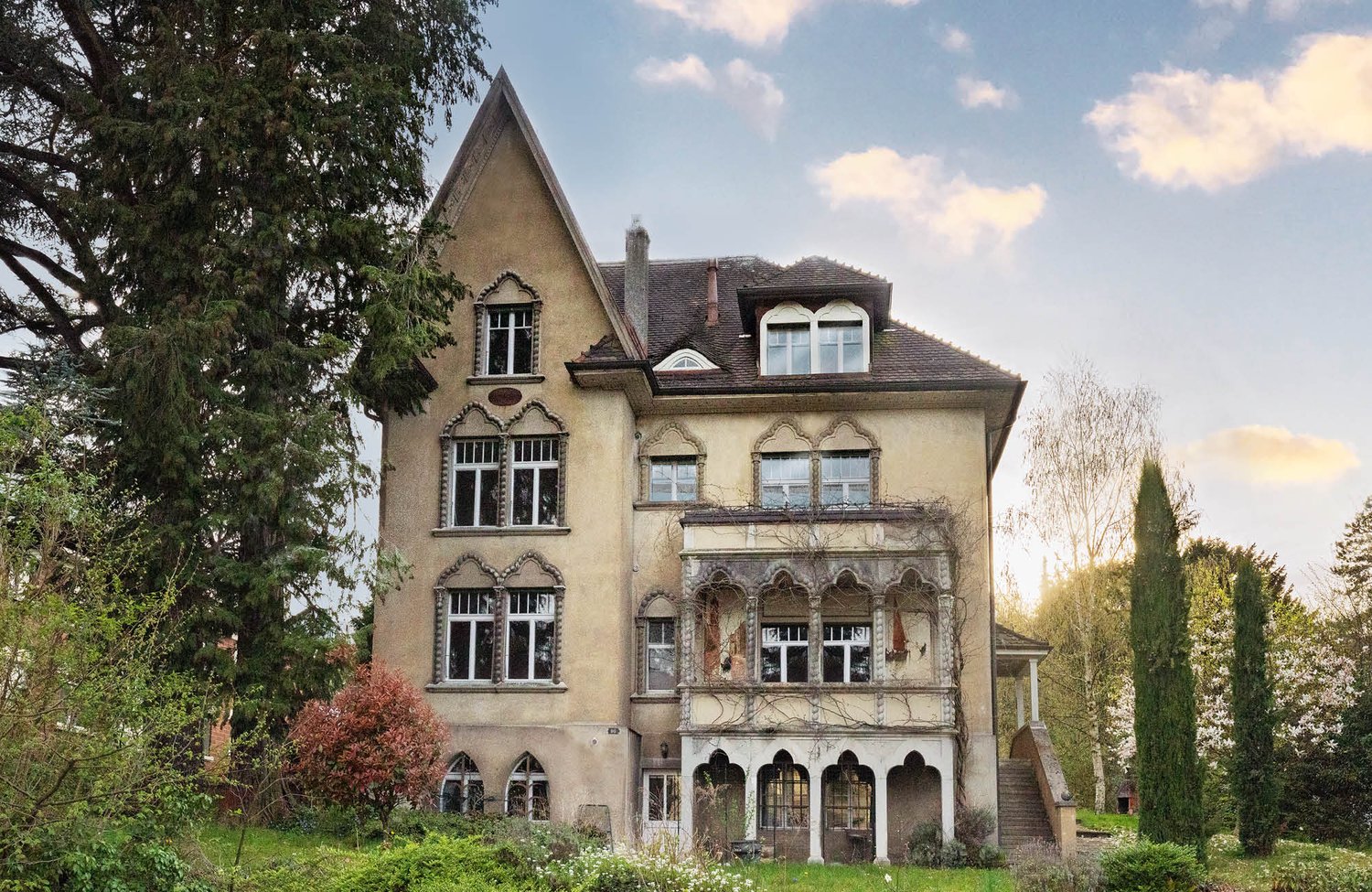 A large, ornate, multi-story house with a distinctive architectural style featuring arched windows, decorative elements, and a sloped roof. The house is surrounded by trees and greenery, giving it a picturesque, historic appearance.