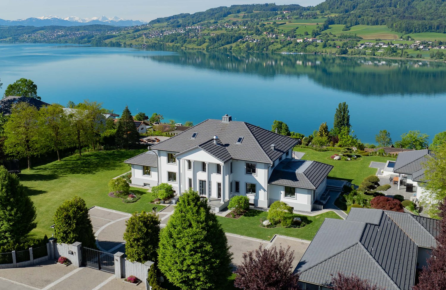 Large two-story white house with a slate roof, surrounded by a well-manicured lawn and gardens. The house has multiple balconies and a garage. It is situated on a hill overlooking a scenic lake and mountains in the background.