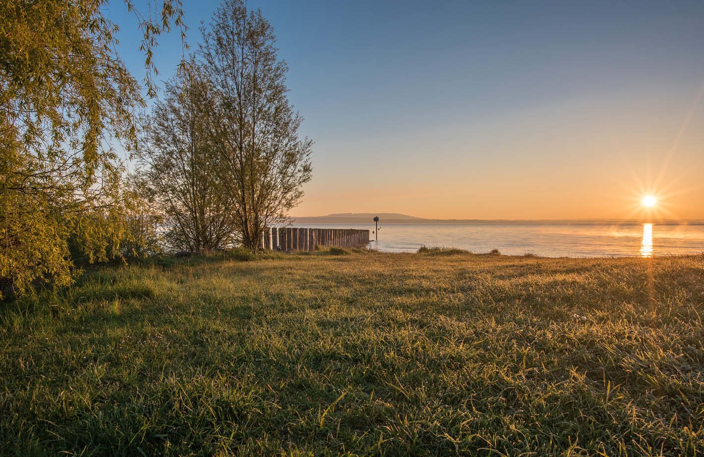 The image shows a scenic lakeside landscape at sunset. There is a grassy field in the foreground, with trees lining the shore. In the distance, the lake can be seen with mountains on the opposite shore. The sky is filled with warm, golden hues as the sun