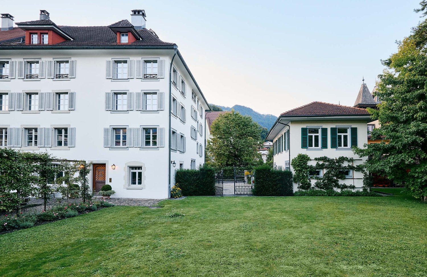 2 story white building, windows with shutters, brown roofs, garden with bushes, green grass, mountains in the distance