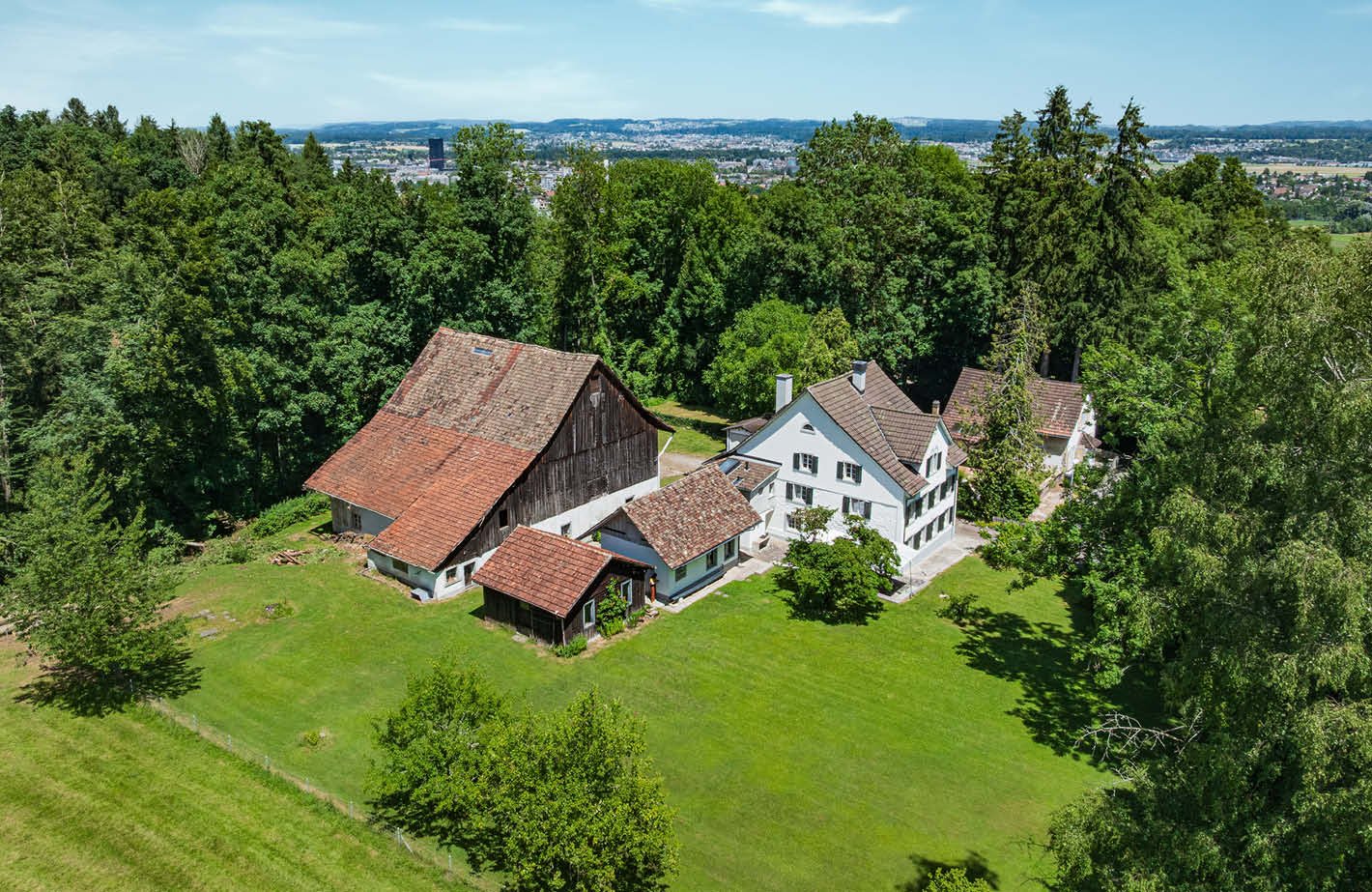 Two-story farmhouse with a red tile roof, surrounded by lush green trees and a large grassy lawn. The property also includes a smaller wooden barn-like structure. The houses have white walls and windows, and there appears to be a balcony or terrace on one