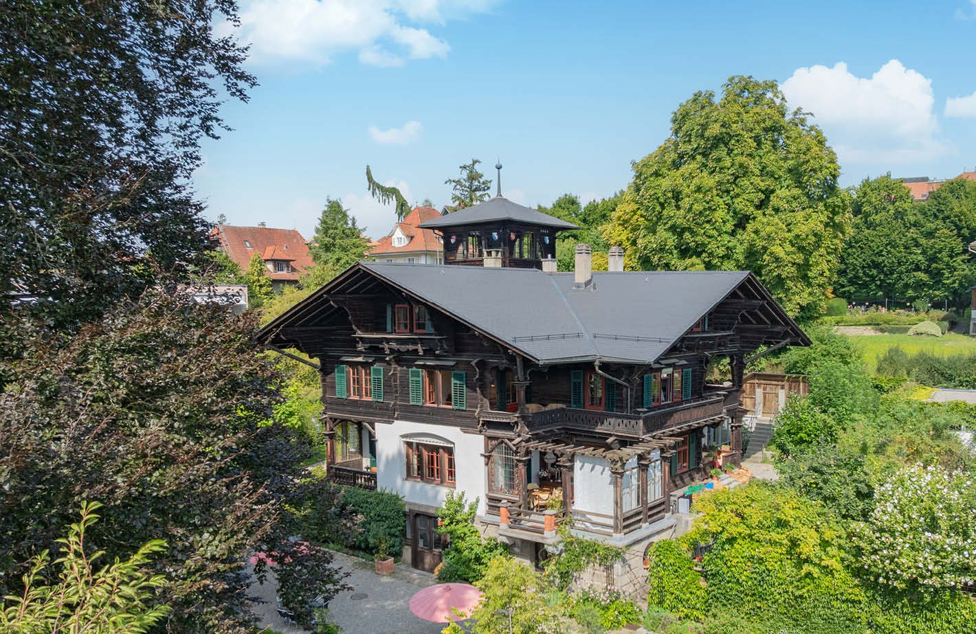 A large 2 story chalet style house with a gray roof, a small tower, green shutters, and a balcony.