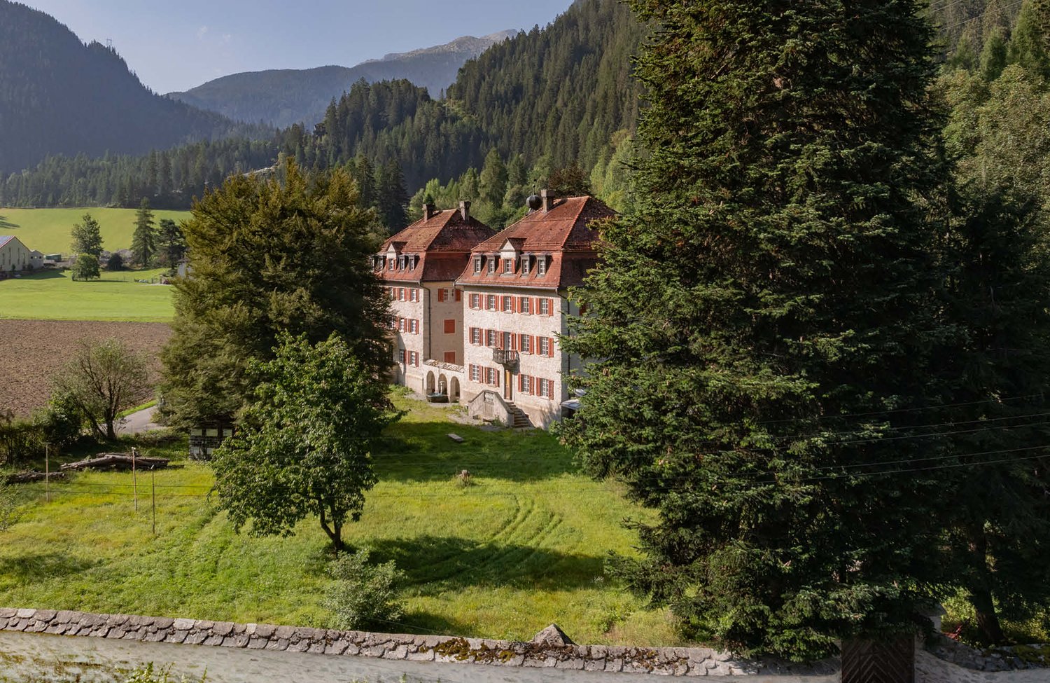 Large historic manor house with red-tiled roofs surrounded by lush green forests and mountains in the background. The house appears to be situated on a grassy field with a stone wall in the foreground.