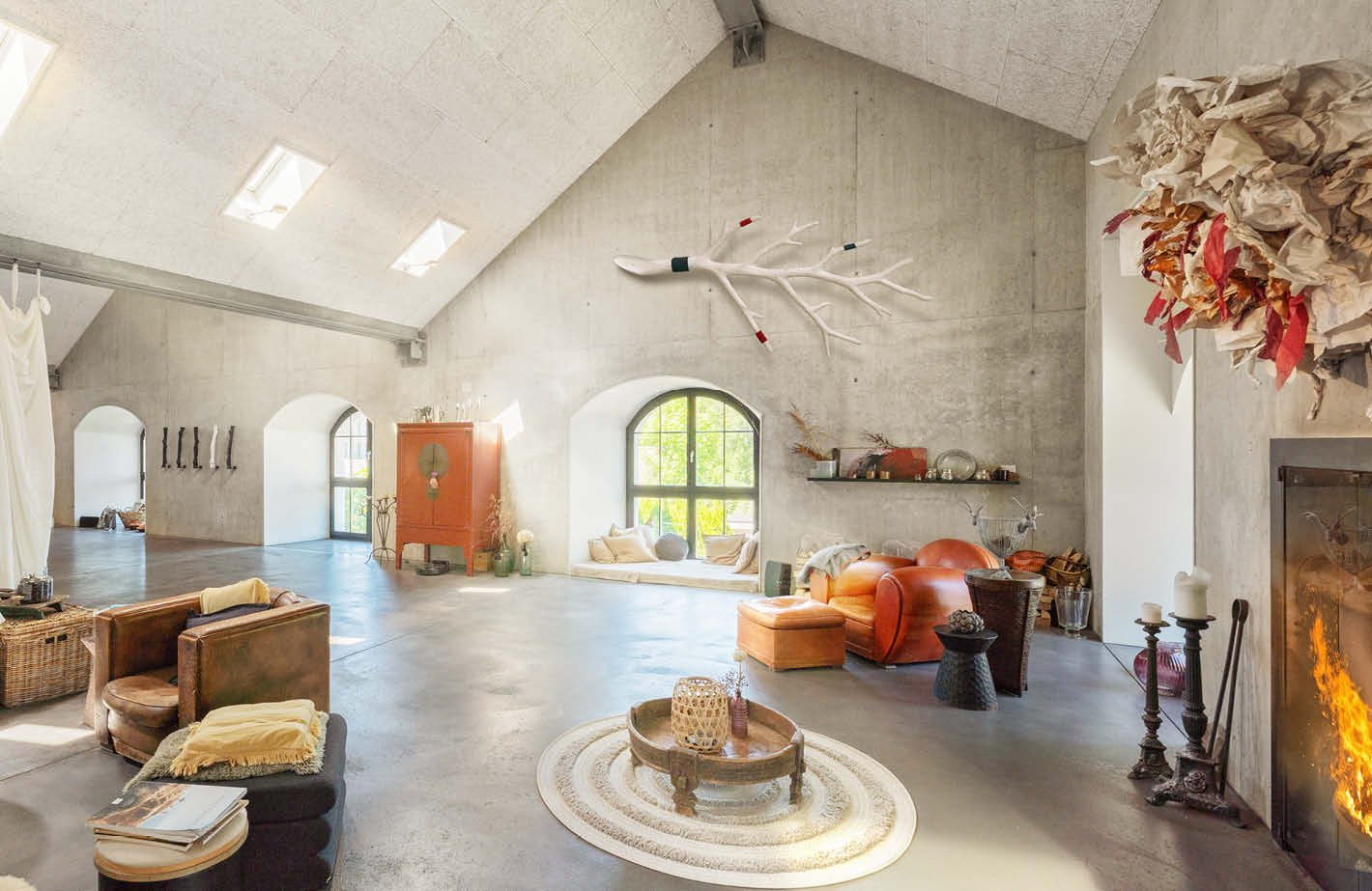 Spacious open-concept living area with high ceilings, arched windows, concrete walls, and a fireplace. The room features a mix of rustic and modern decor elements, including a large orange sofa, wicker furniture, and decorative accents like a hanging plan