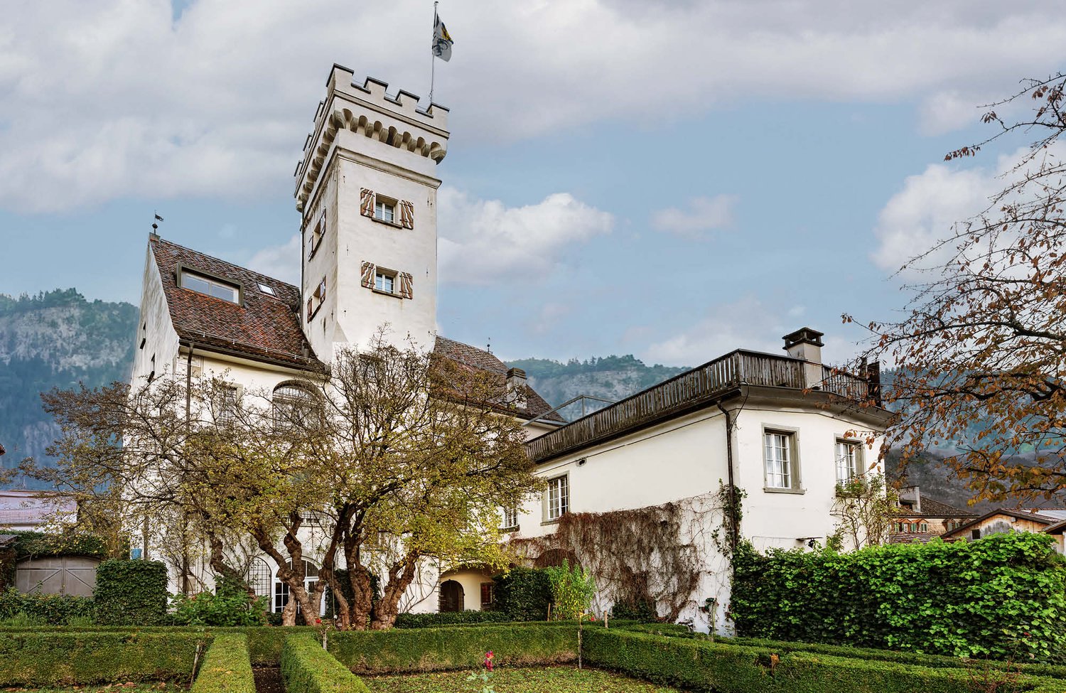 Large white house with tower, flag, red roof, garden, mountains in the background