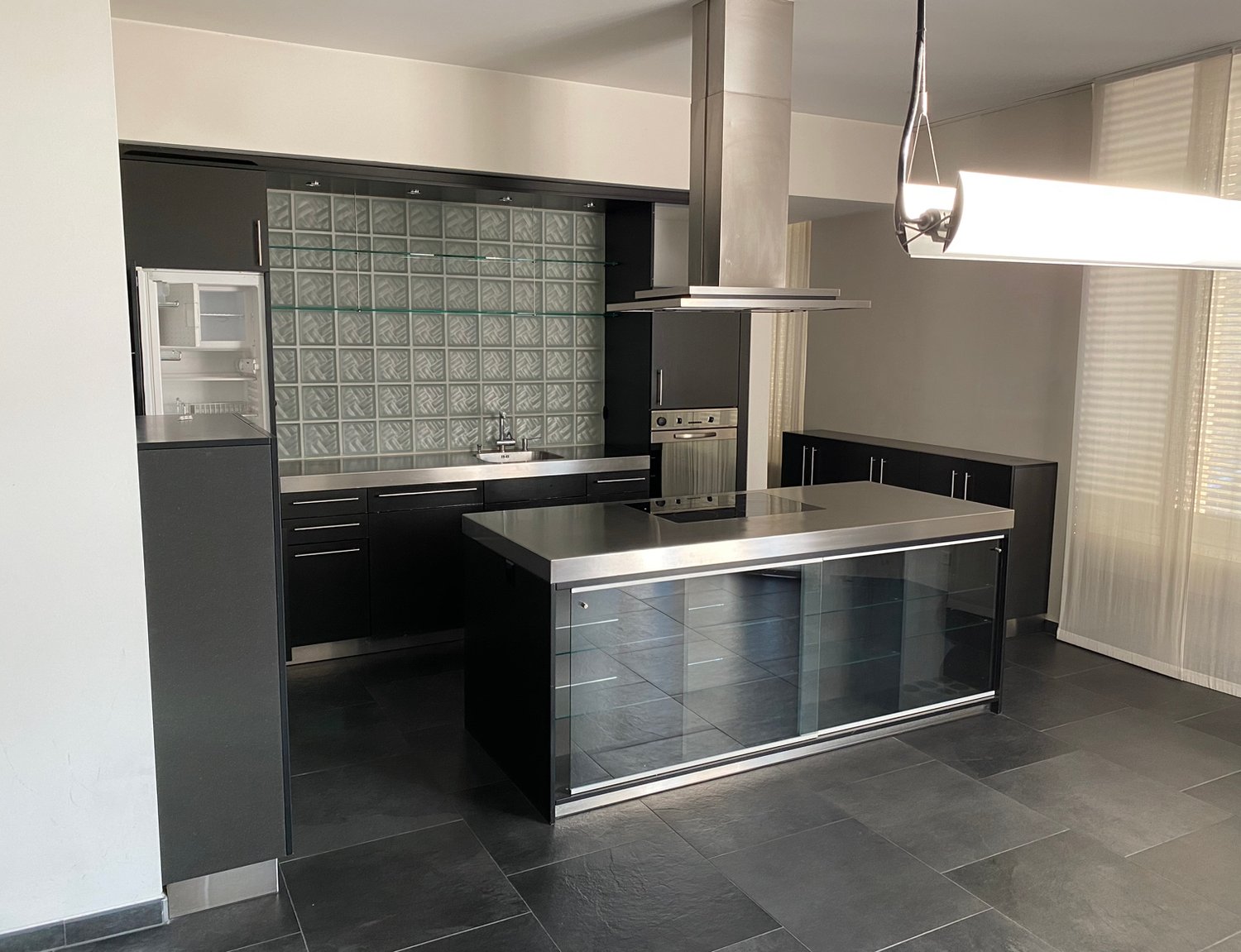 Modern kitchen with island, stainless steel countertops, black cabinets, glass wall, central stove, refrigerator