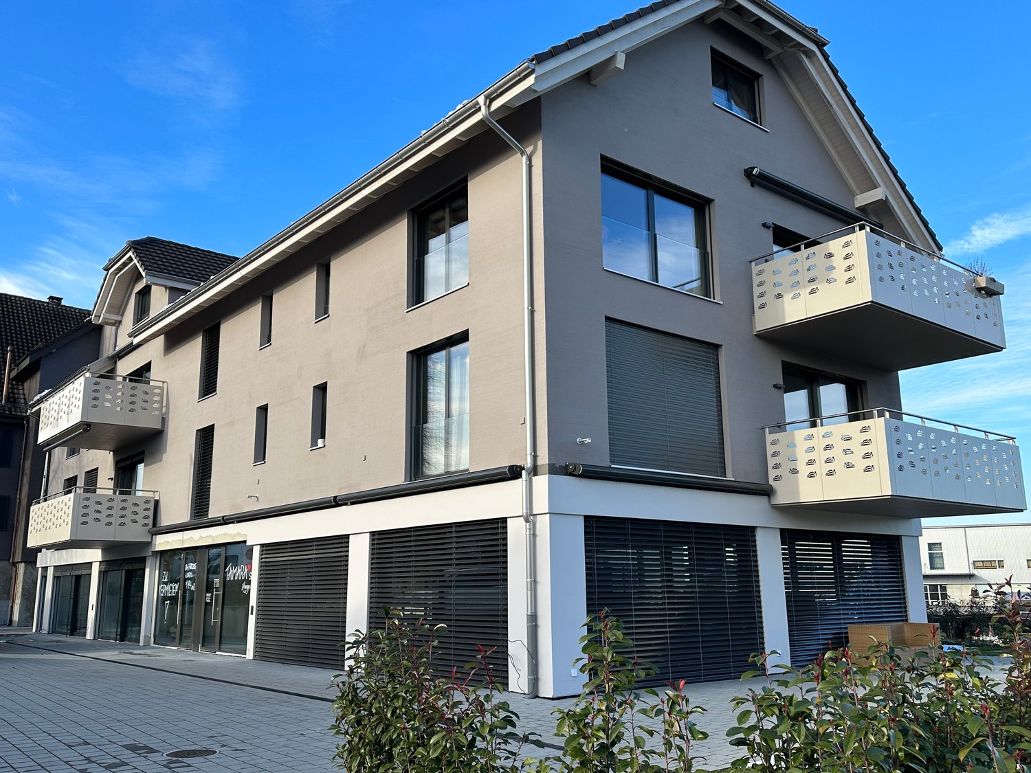 Modern building, concrete wall, grey color, 4 floors, many balconies, glass windows, sliding glass doors, ground floor shops, street in front.