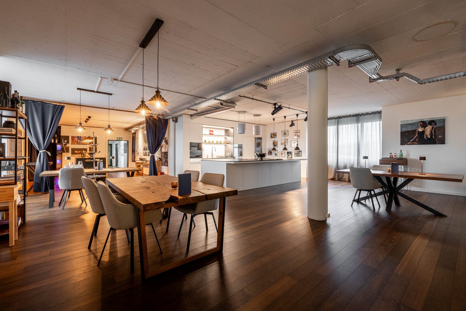 Dining area with wooden tables, chairs, and a kitchen island. The room has wooden floors and white walls. There are lamps hanging from the ceiling and a painting on the wall.