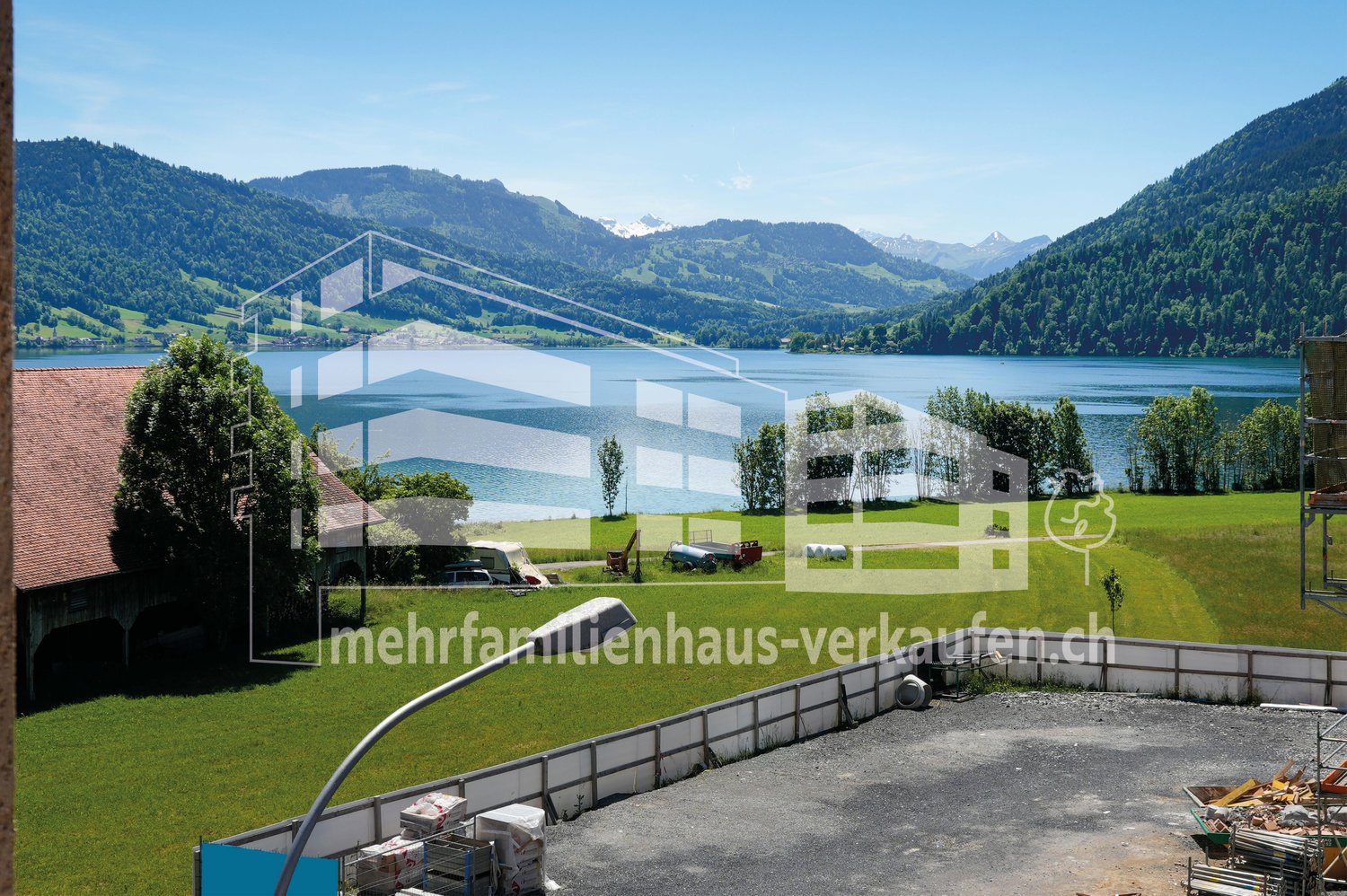 House with a view of a lake and mountains, surrounded by a green field, with a barn and a building in the background.
