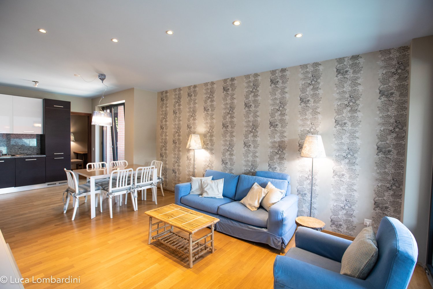 wooden floor, blue couch, wooden table, wooden coffee table, dining table, lamps, kitchen visible