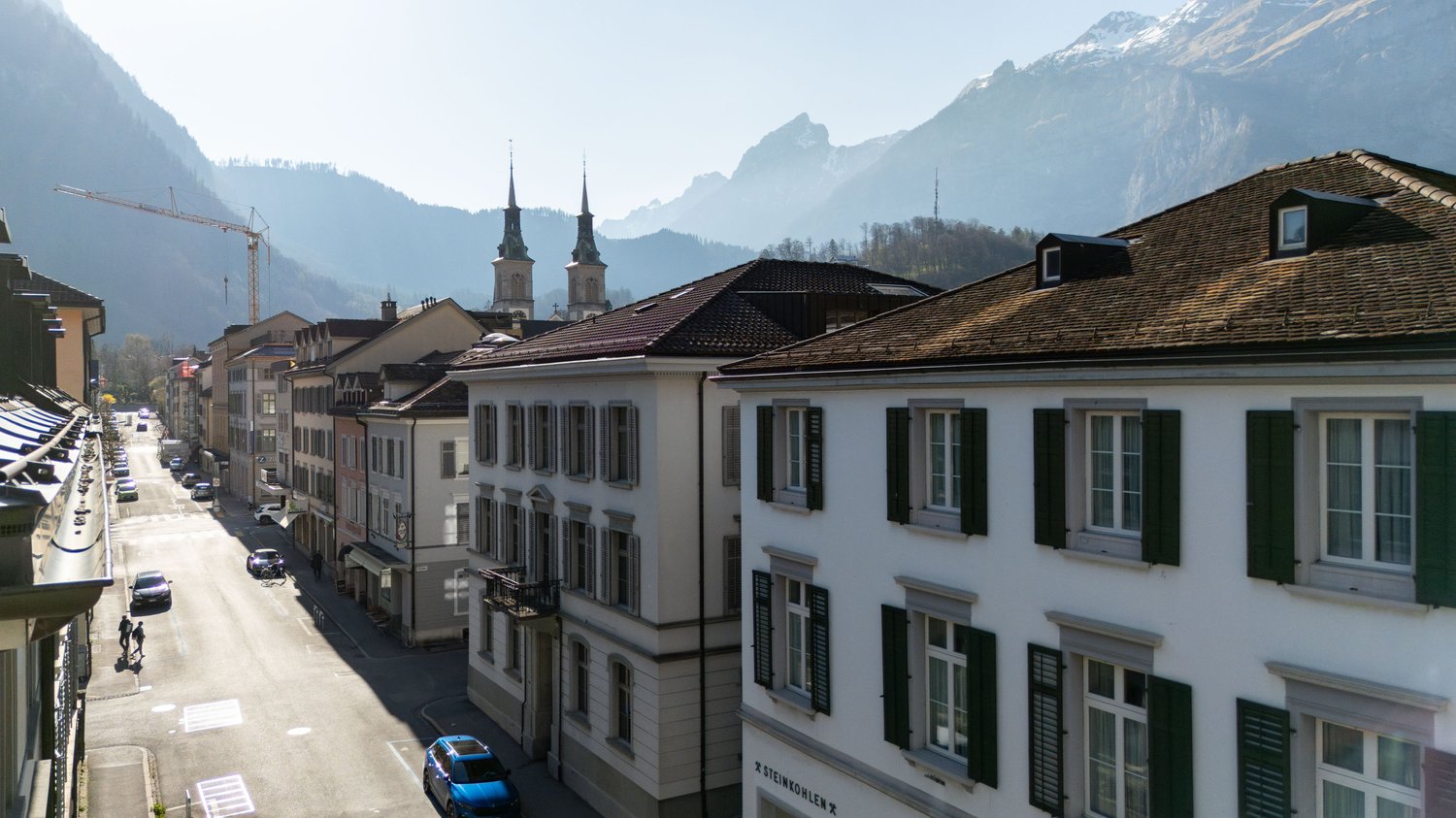 This image shows a picturesque street in a European city, with traditional buildings featuring white facades, black shutters, and red tile roofs. In the background, snow-capped mountains can be seen, creating a stunning natural backdrop. The street is lin
