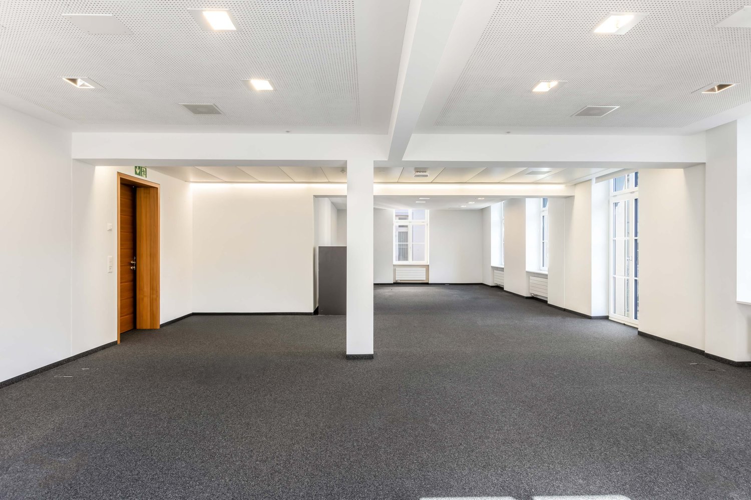 An empty office space with white walls, gray carpet, and a wooden door on the left side.