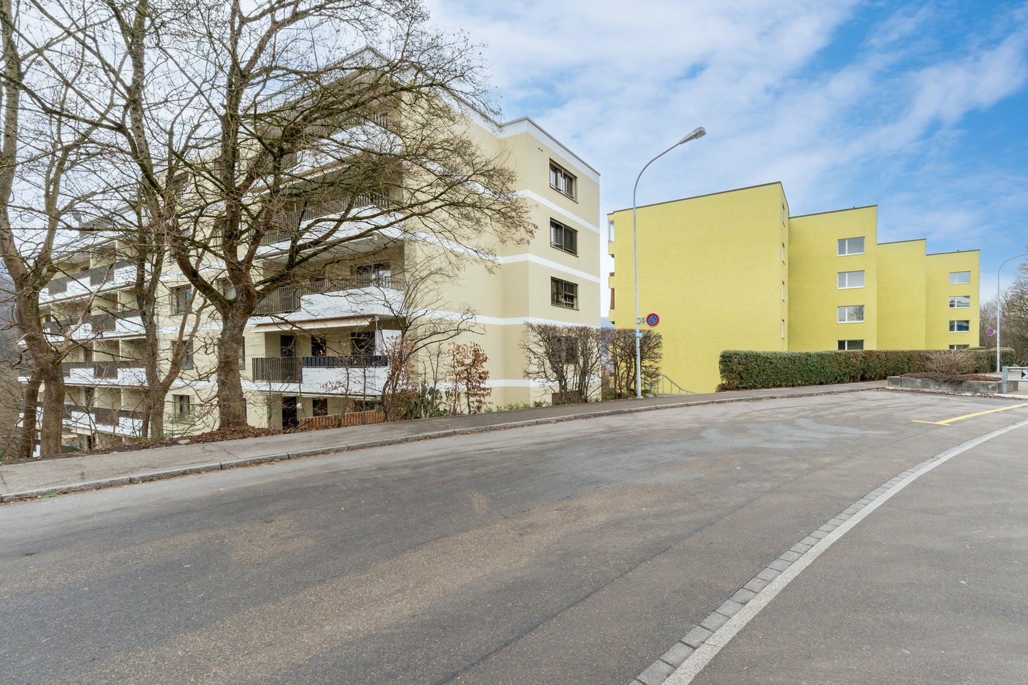 Large multi-story building with balconies, beige exterior, modern design, surrounded by trees and bushes, clear sky