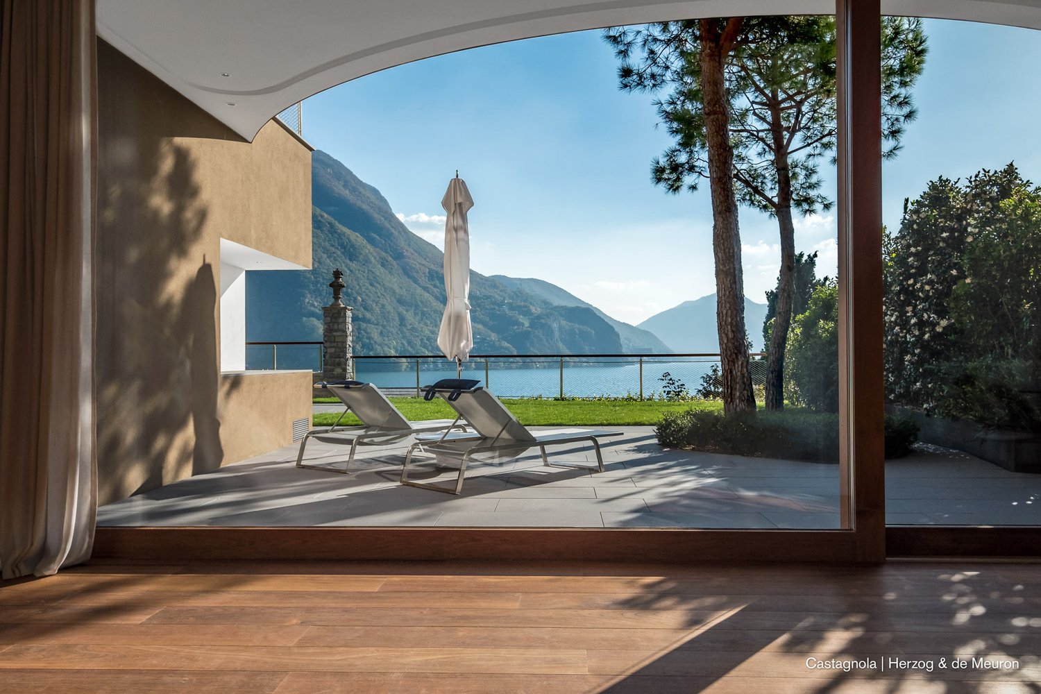 large terrace with lounge chairs and umbrella, wooden floor, large windows, mountains and lake view