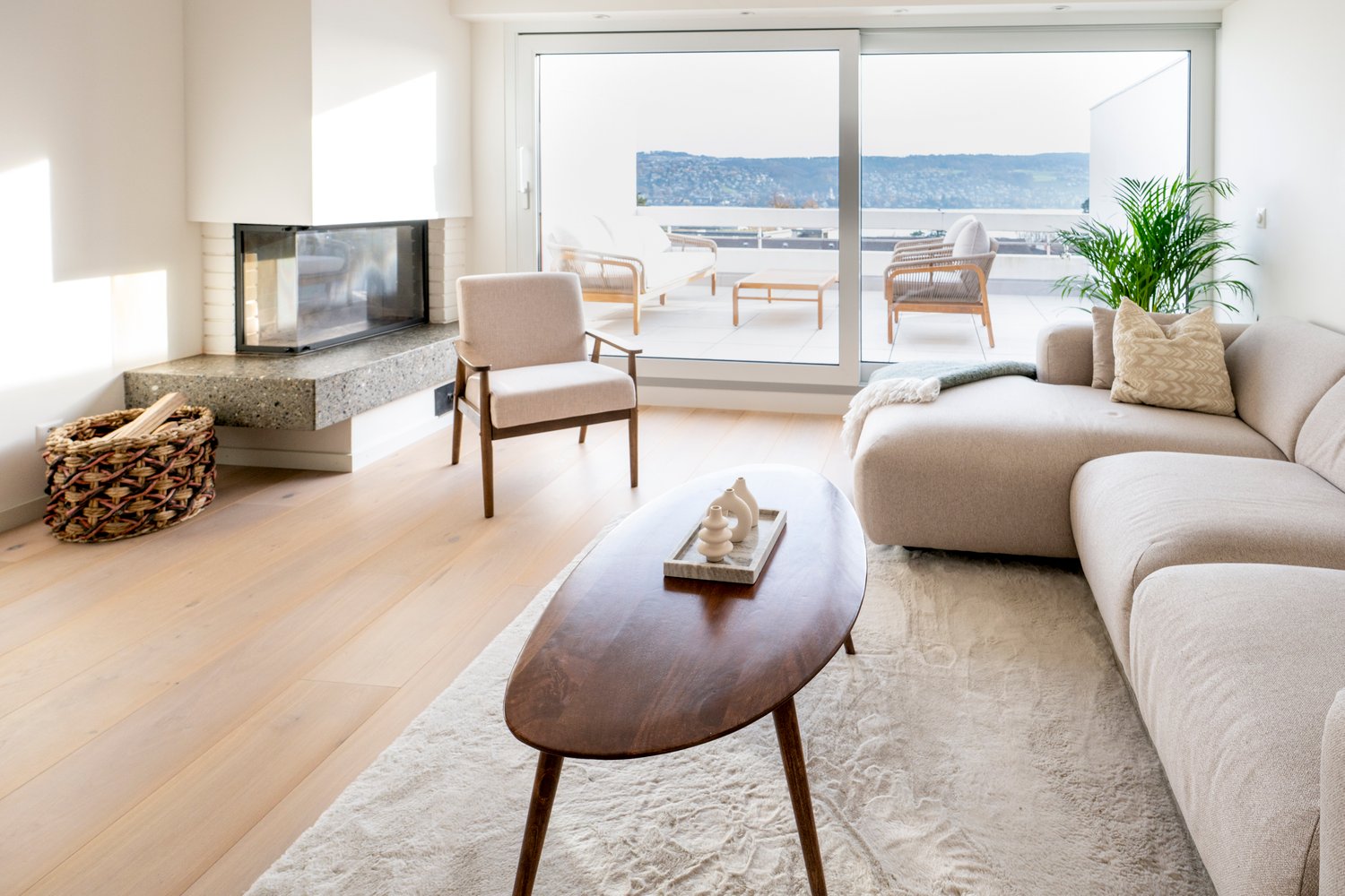 White walls, hardwood floors, fireplace, a couch with pillows, an oval coffee table, large windows leading to a balcony, a wicker basket with firewood, a plant, and a view of the mountains