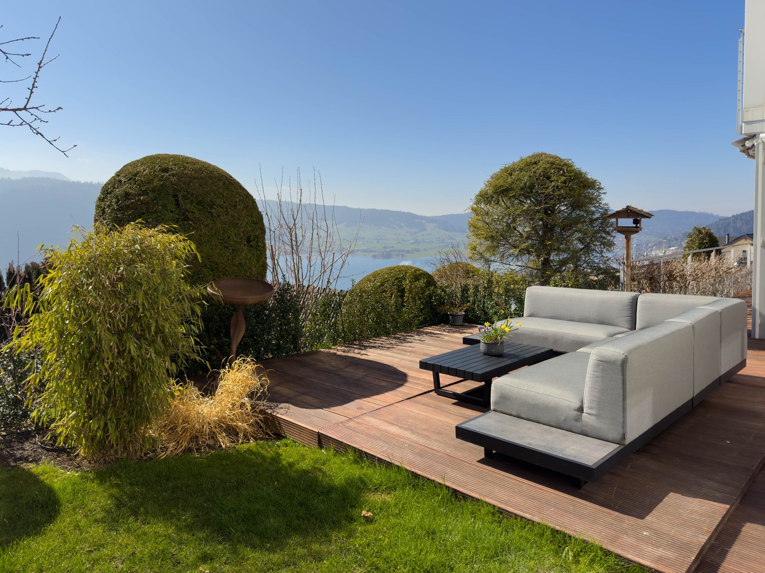 Wooden deck with comfortable outdoor seating, surrounded by lush greenery and landscaping, with a scenic view of the mountains and lake in the background.