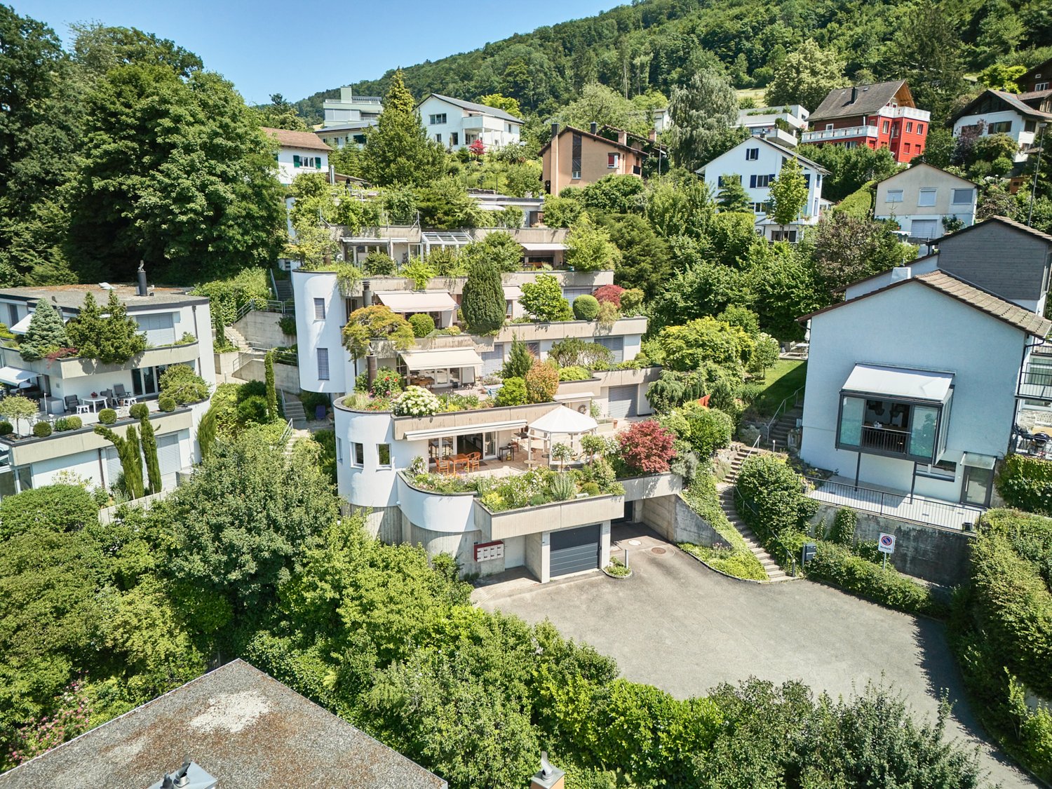 The image shows a residential neighborhood nestled in a lush, green hillside. The houses have various architectural styles, with some featuring balconies and terraces. There are also parking spaces visible in the foreground, indicating the presence of gar
